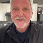 A man with a beard and a black jacket is smiling in a kitchen.