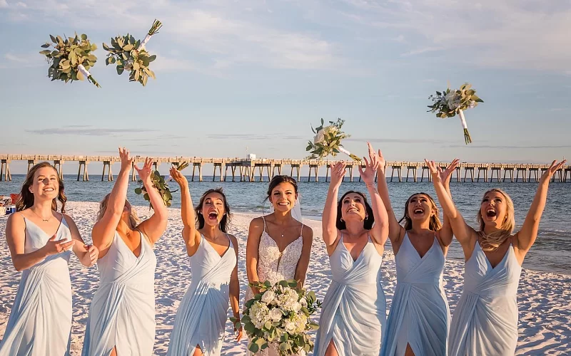 A bride and her bridesmaids are throwing their bouquets in the air on the beach.