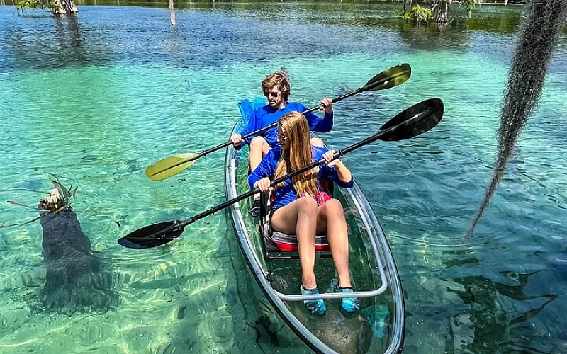 A boy and a girl are paddling a clear kayak on a lake.