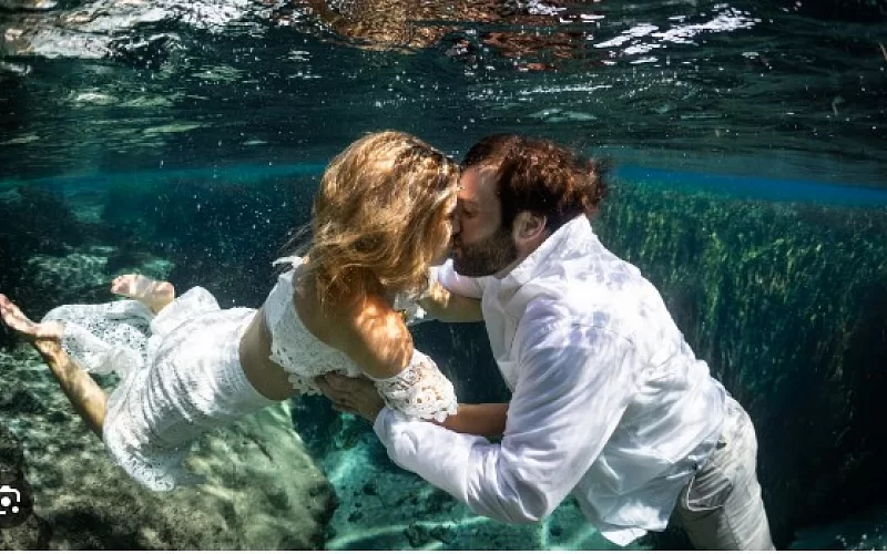A bride and groom are kissing underwater in a pool.