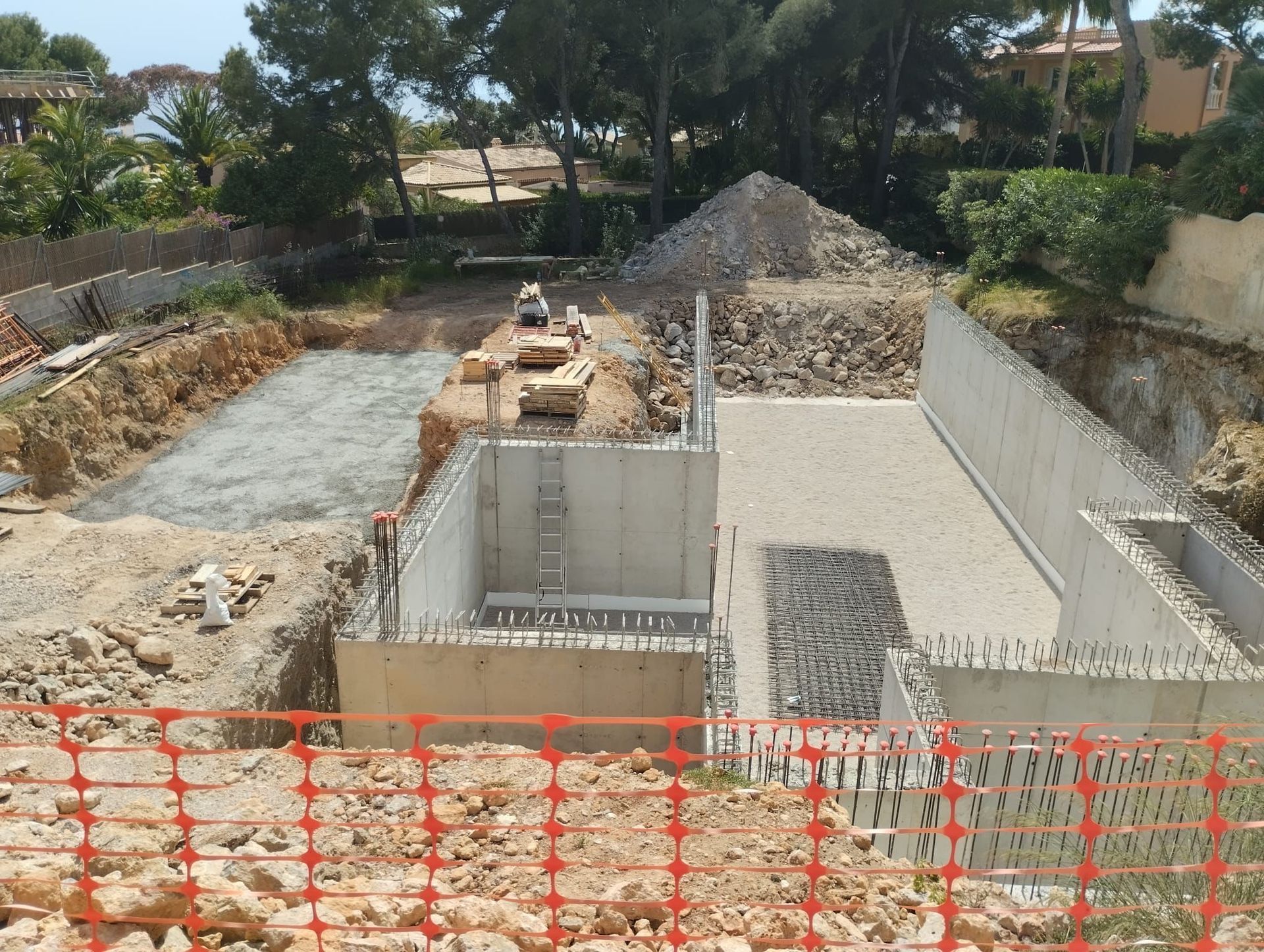 Obra en construcción: muros de hormigón, barras de refuerzo, movimiento de tierras, valla de seguridad naranja.