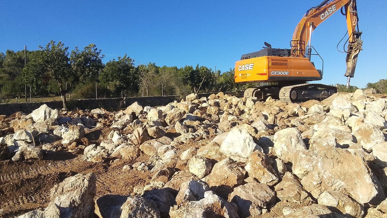 Una excavadora CAN RAMIS rompe rocas en un campo de tierra bajo un cielo azul brillante.