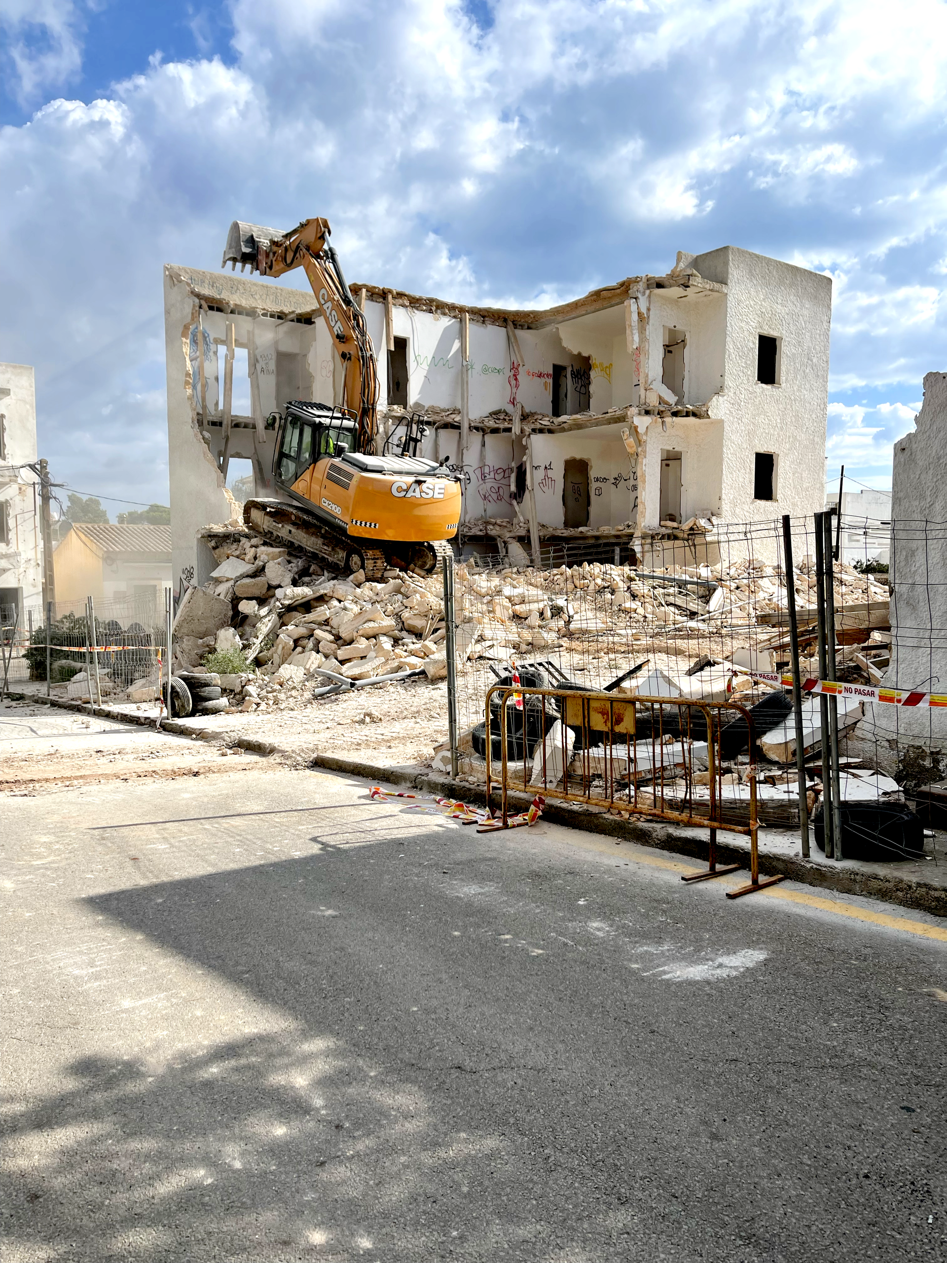 Una excavadora CAN RAMIS demuele un edificio blanco bajo un cielo nublado. Los escombros cubren el suelo.