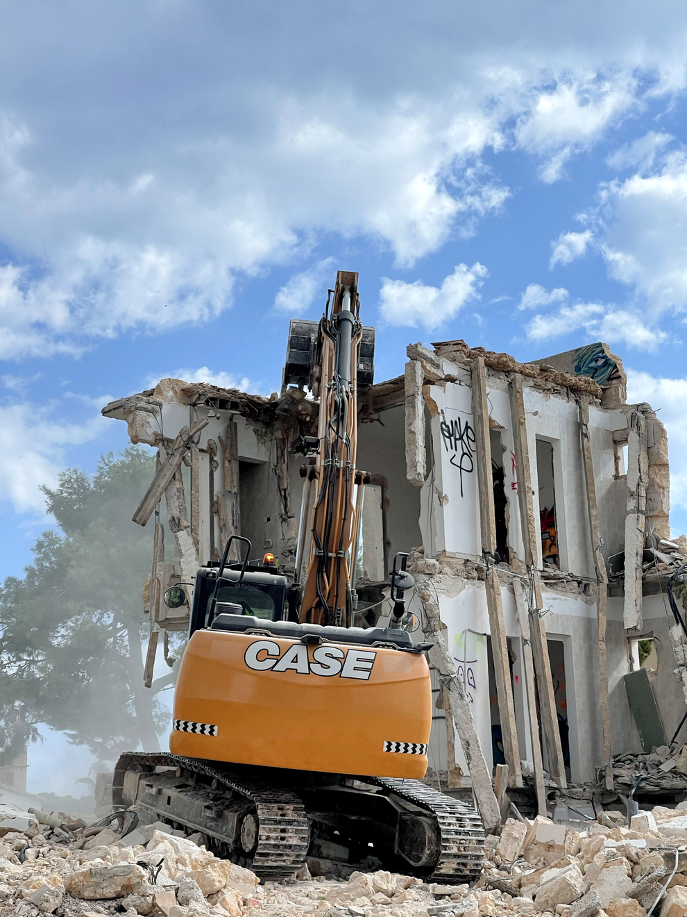 Excavadora demoliendo un edificio de dos plantas en un día soleado con cielo azul nublado.