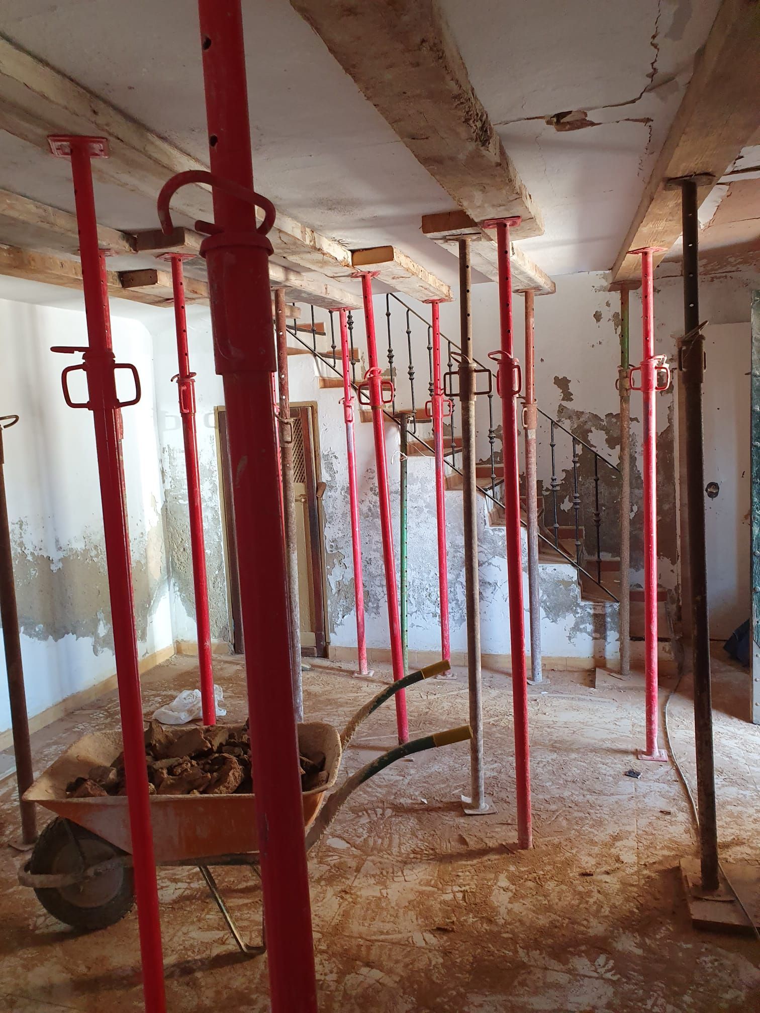 Escenario interior de una obra en construcción con vigas de soporte rojas, vigas de techo a la vista y una carretilla.