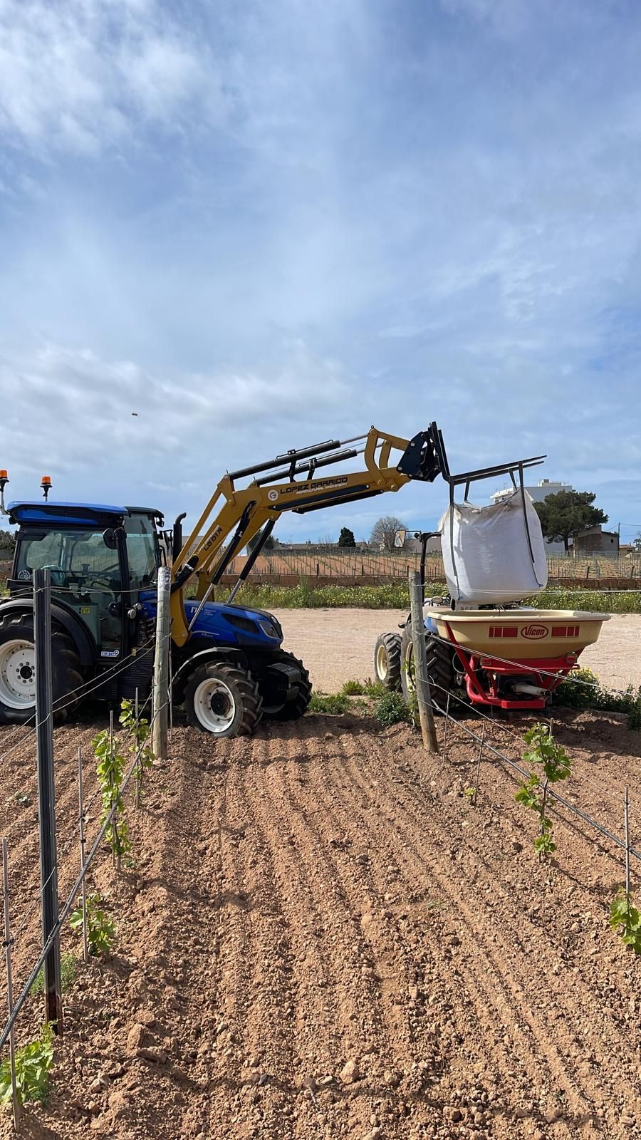 Un tractor con un brazo amarillo levanta un saco grande, distribuyendo fertilizante en un campo.