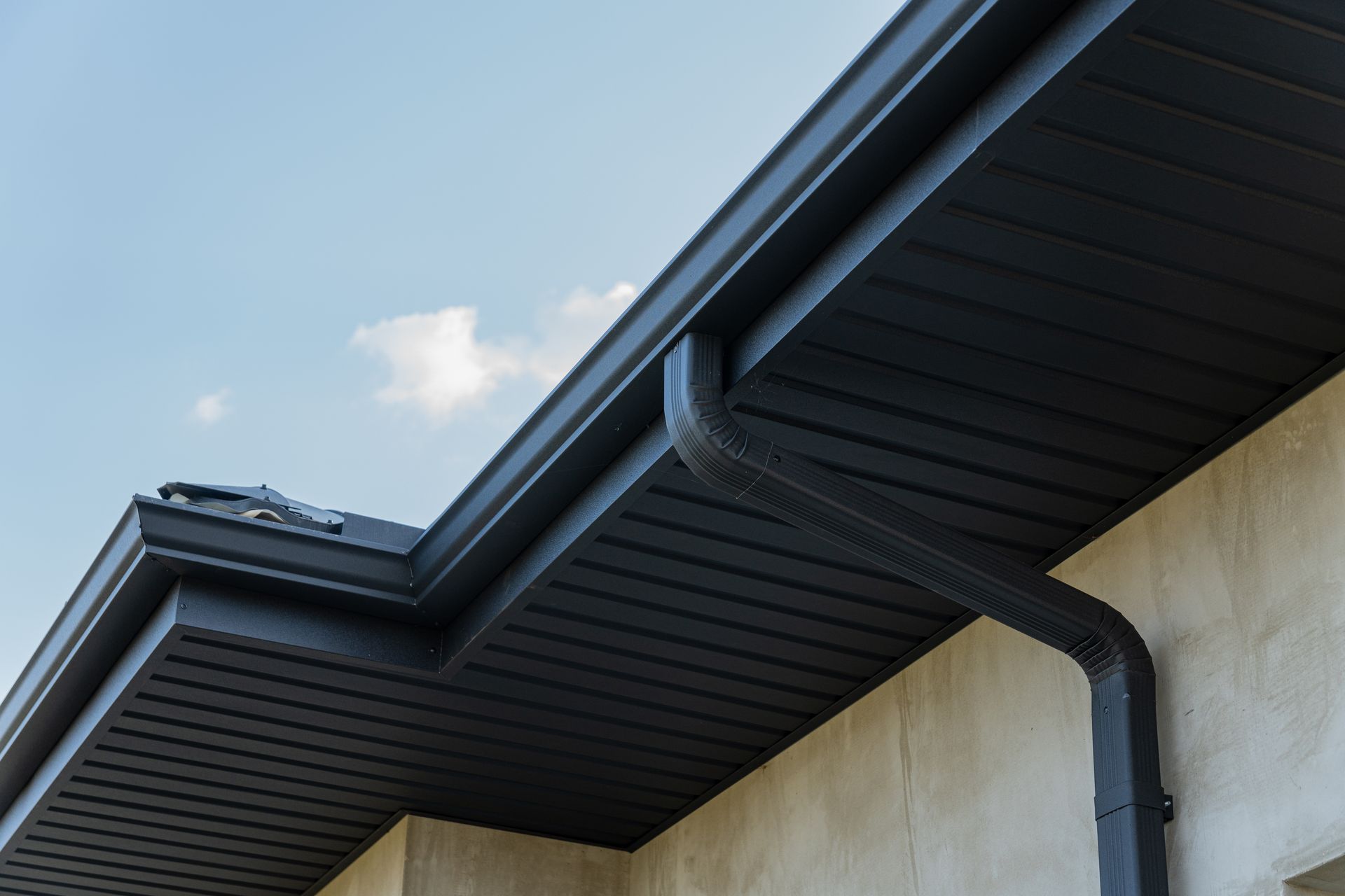 Black rain gutter and fascia against a light blue sky and stucco wall.