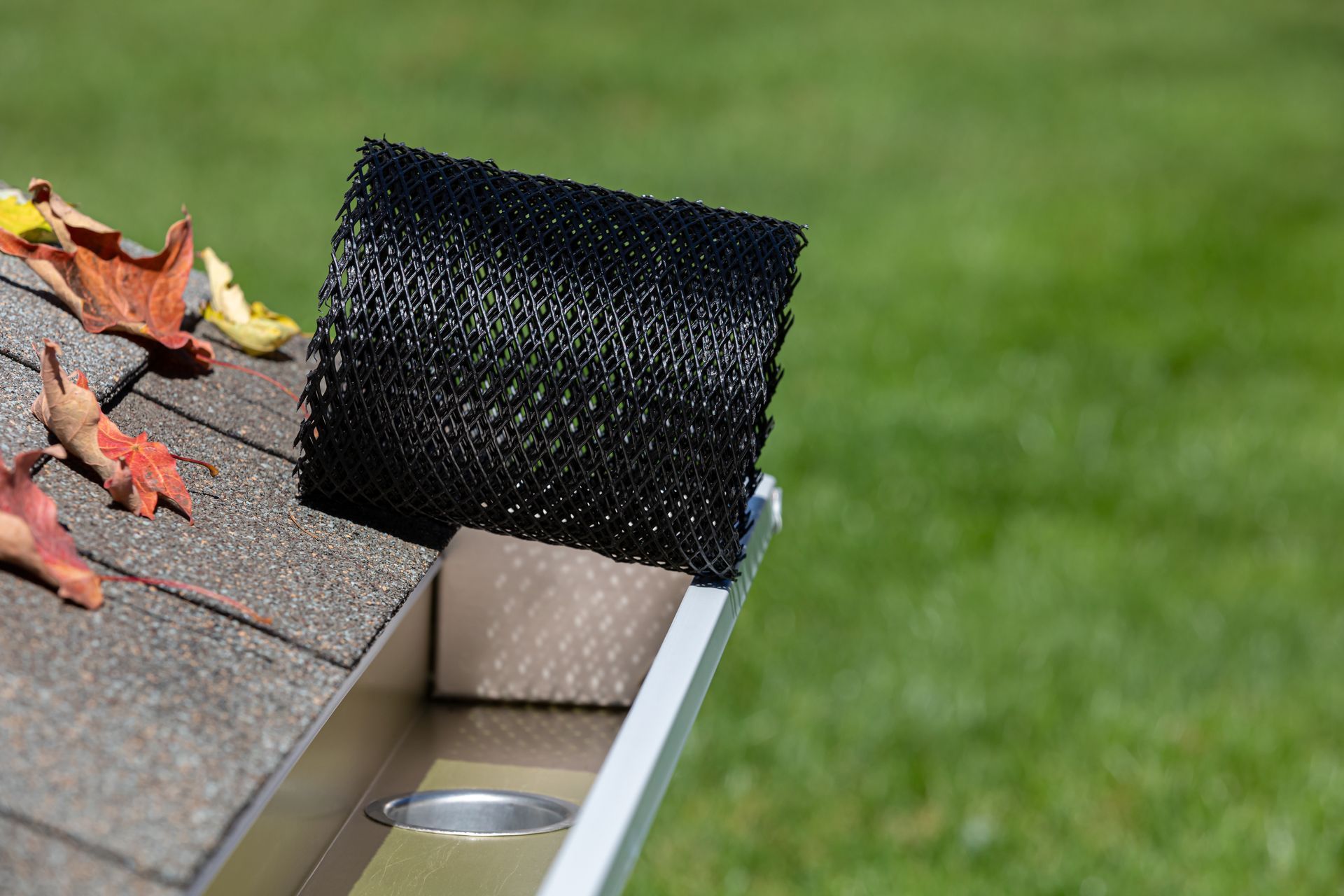 Black gutter guard roll on a roof with leaves, next to a silver gutter and green grass.