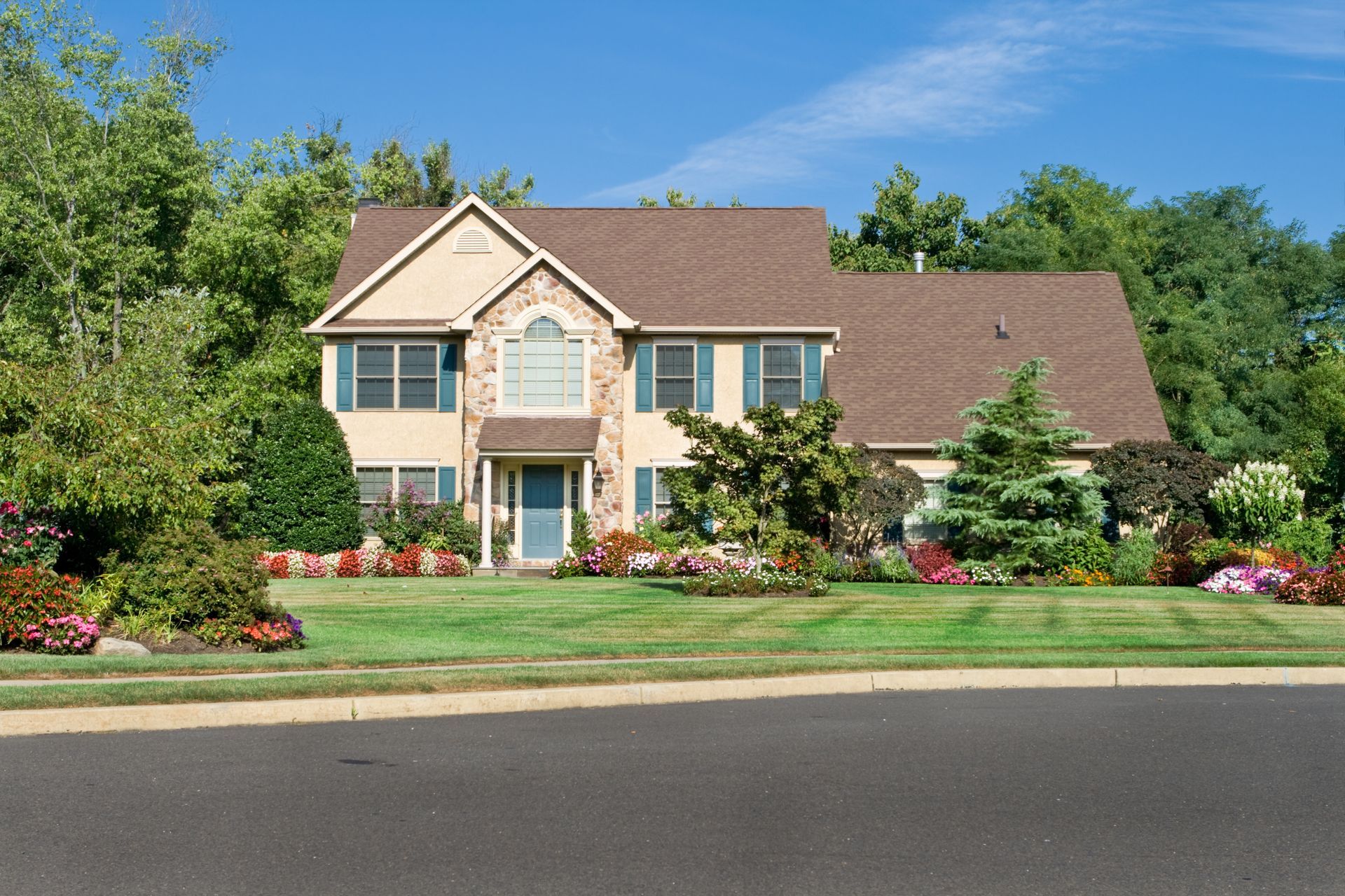 Two-story beige house with brown roof and green shutters, surrounded by green trees and a well-manicured lawn.