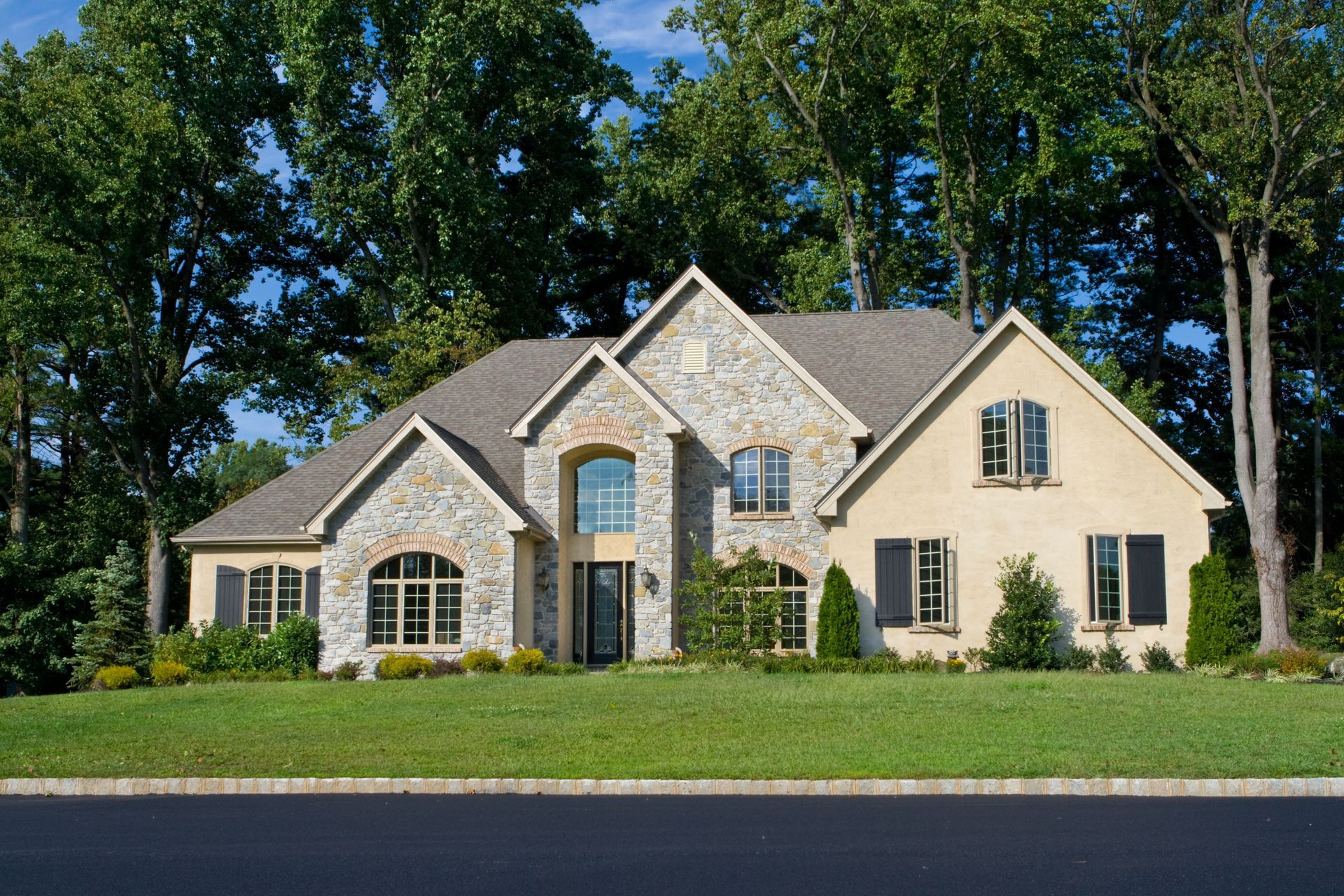 Stone and tan two-story house with black shutters, set in a green yard, with trees in the background.