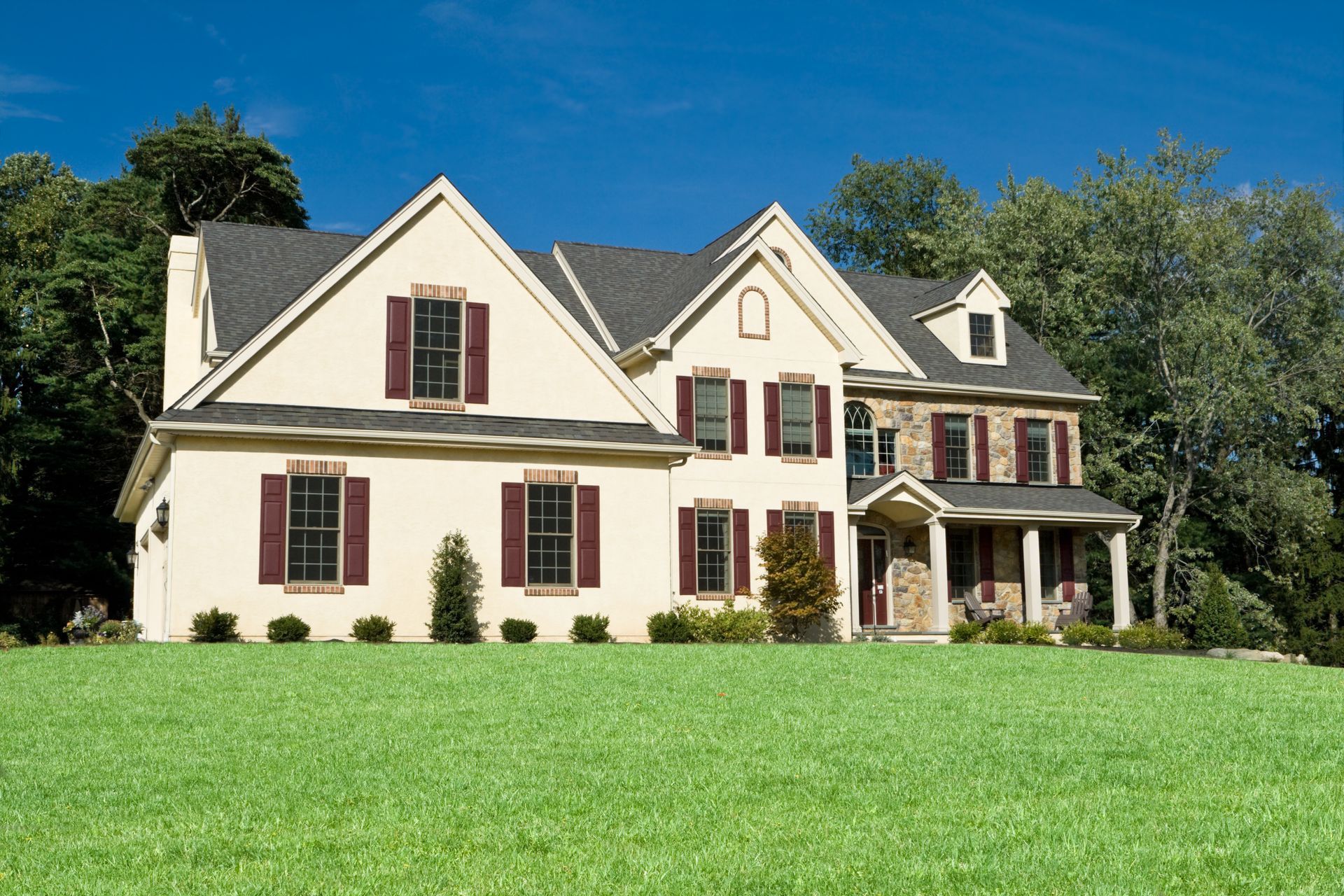Two-story house with tan siding, burgundy shutters, and stone facade, set against green lawn and blue sky.