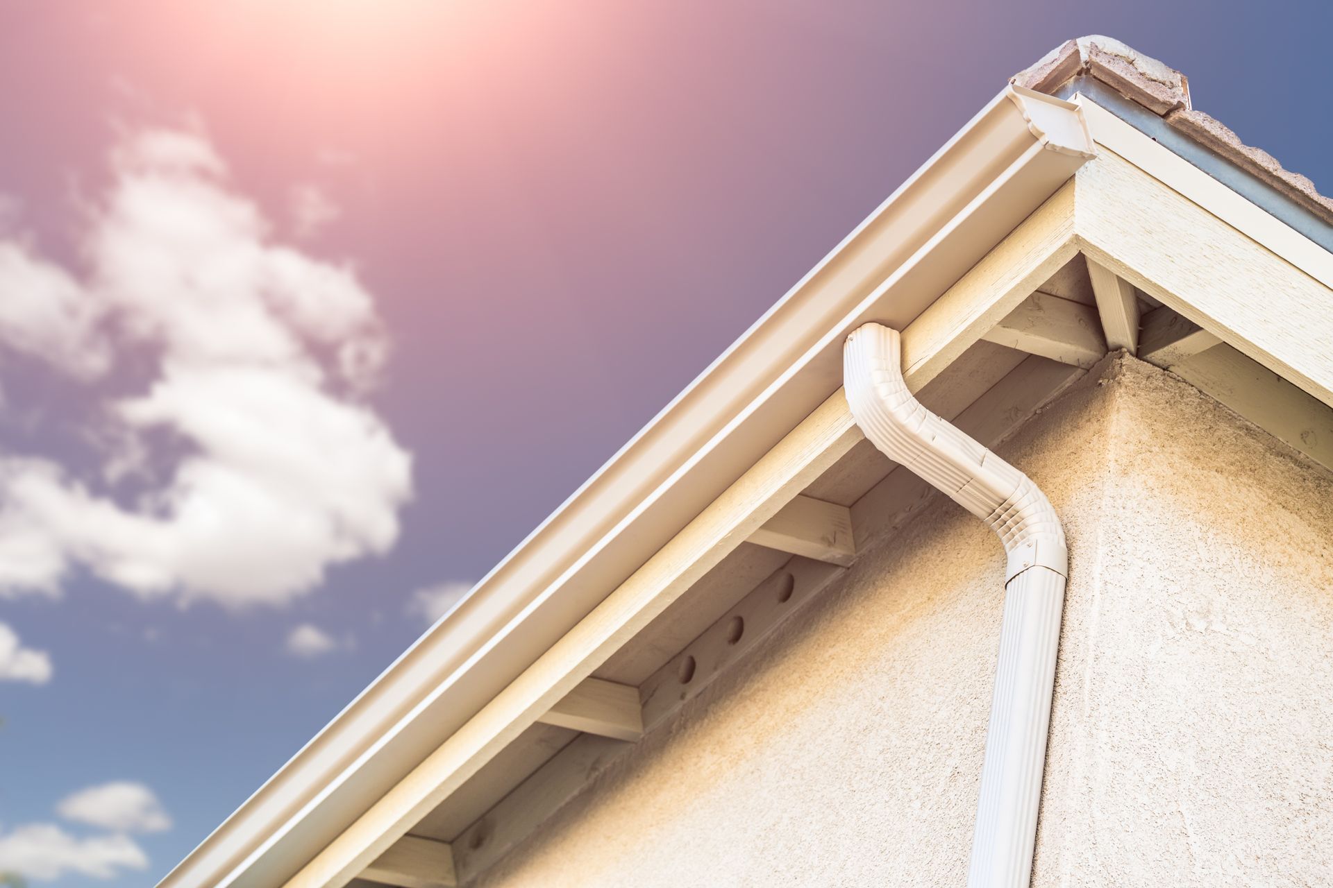 Man in hard hat installing gutter on a red tile roof, against a cloudy sky.