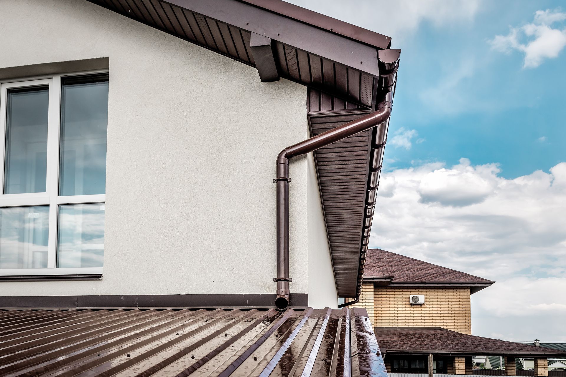 Brown gutter and downspout on a white stucco building with a window and a rooftop, blue sky.