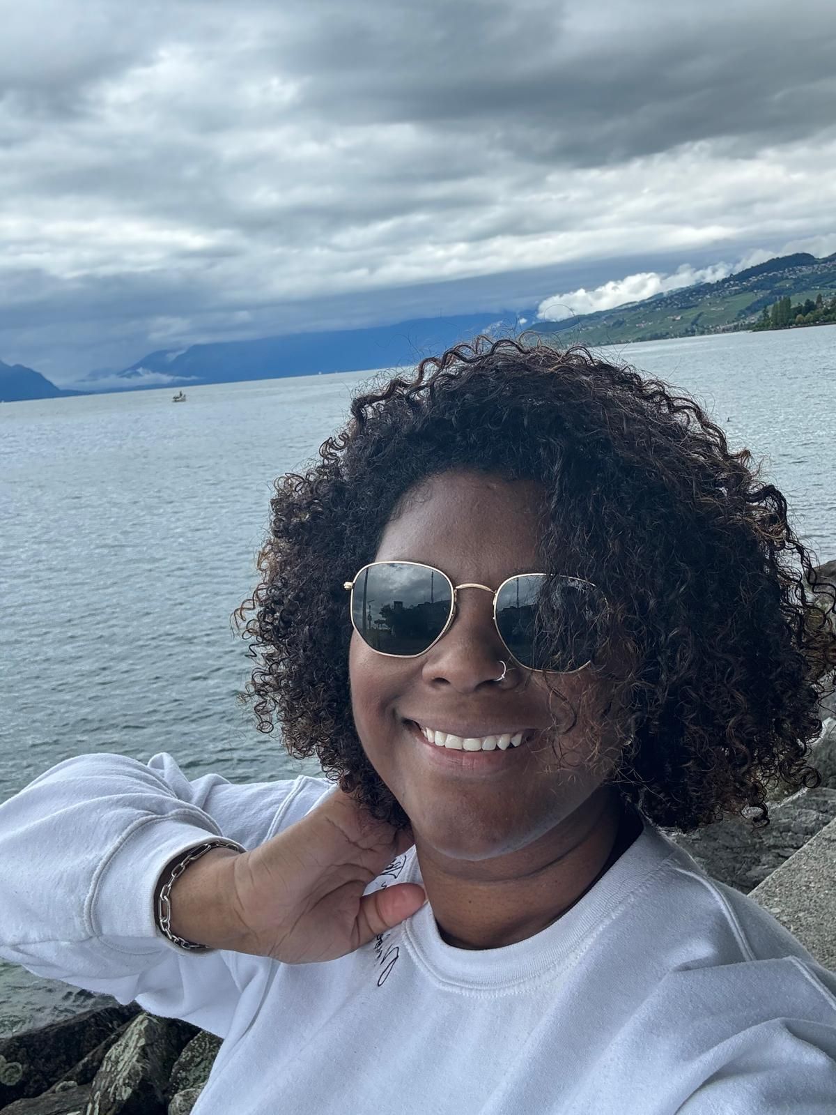 A woman wearing sunglasses is smiling in front of a body of water.