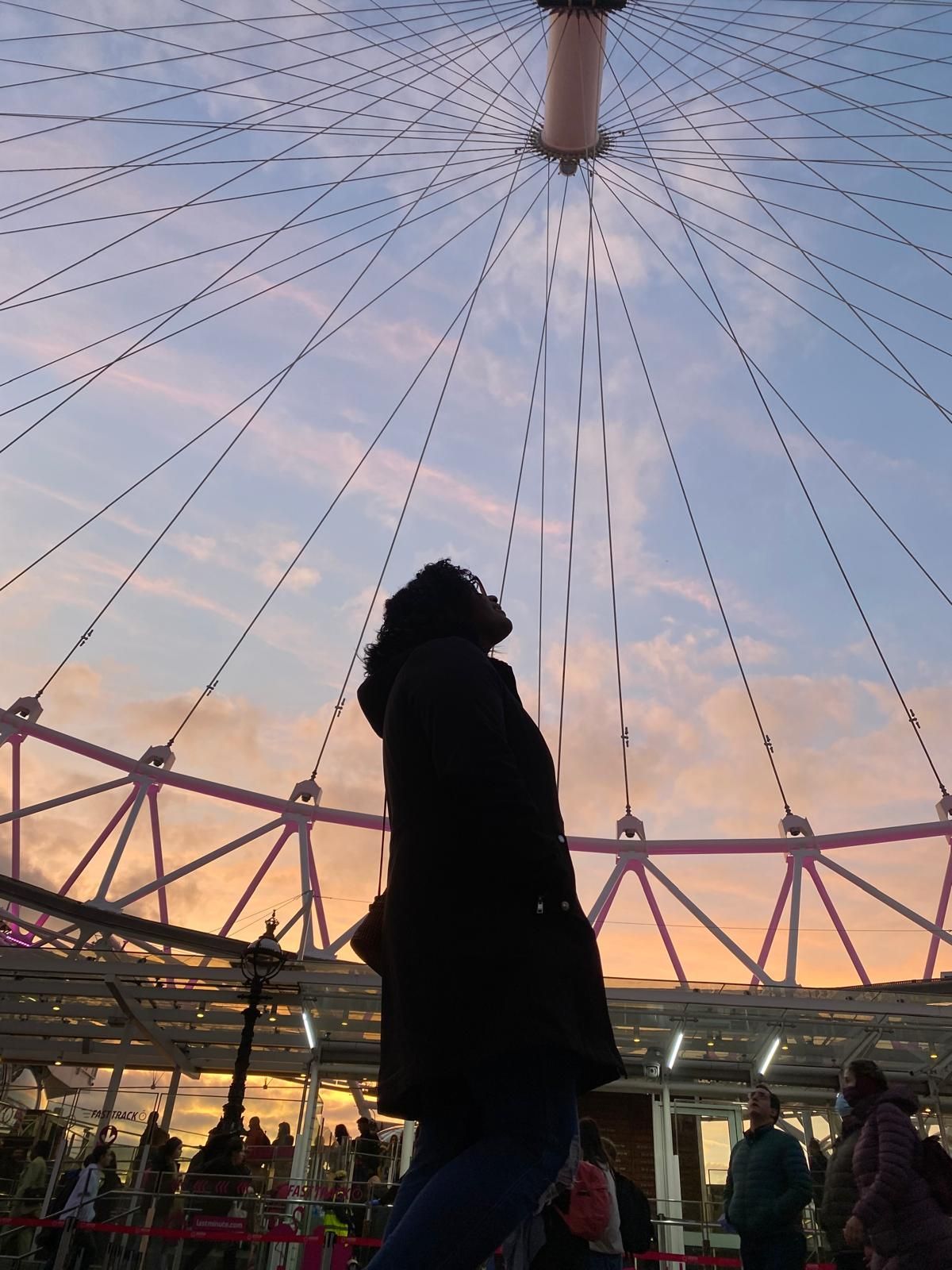 A woman is standing in front of a ferris wheel at sunset.