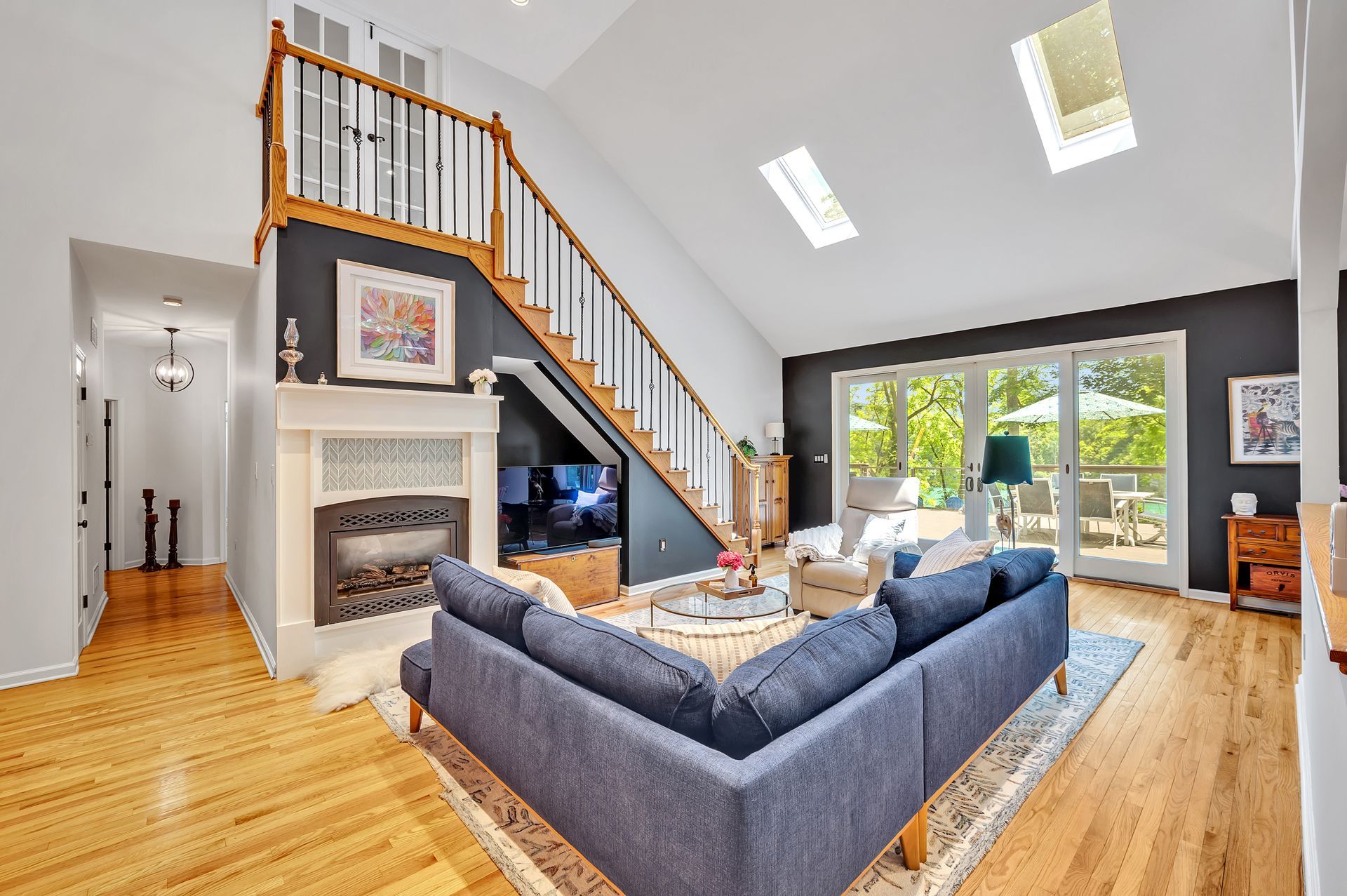 A modern living room with a blue sectional sofa, a fireplace under a wooden staircase, and tall windows with skylights.