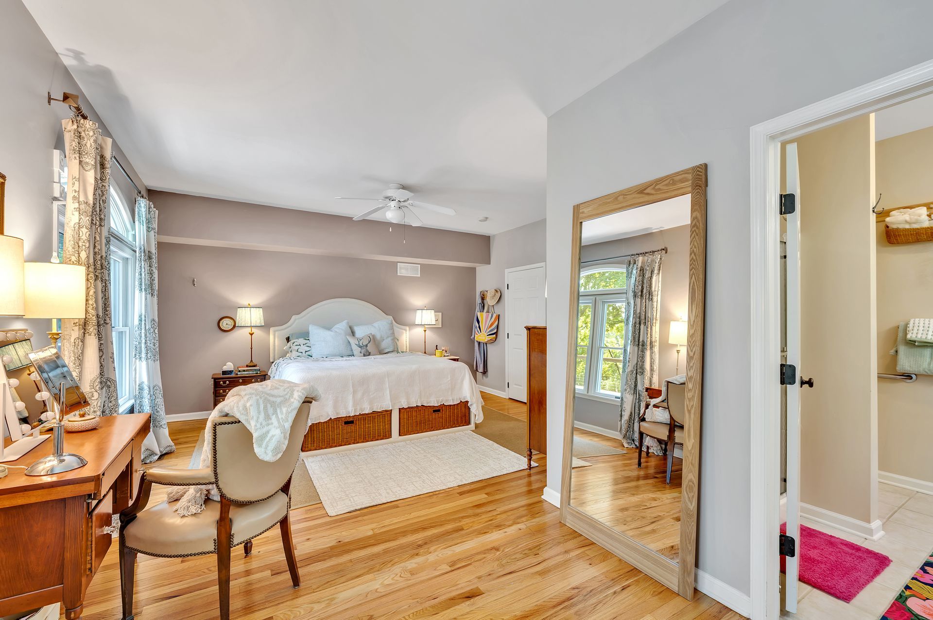 A primary bedroom featuring a king bed, hardwood floors, a wooden desk with a chair, and a full-length floor mirror.