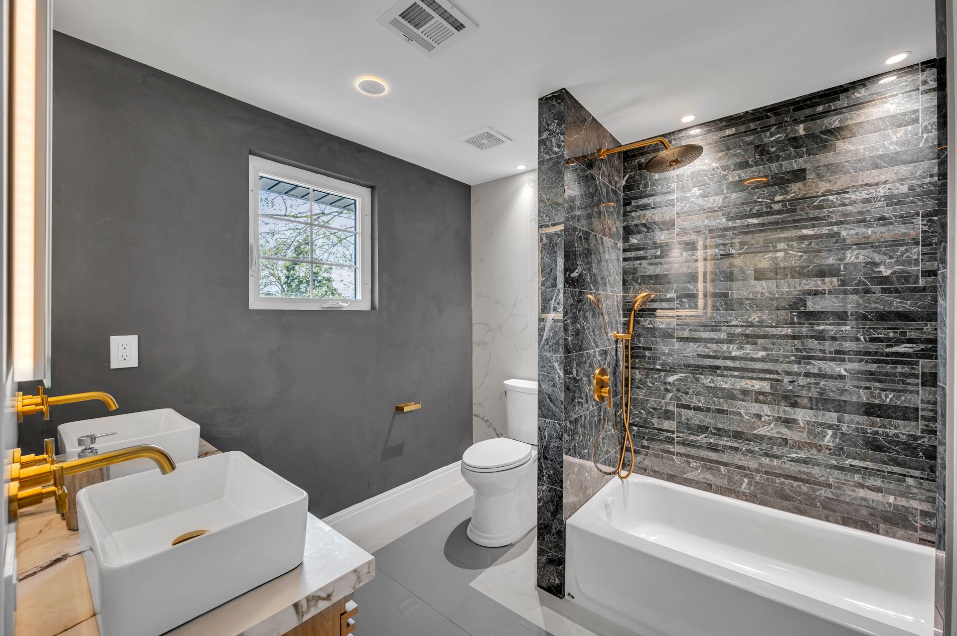 Modern bathroom with dual white vessel sinks, gold fixtures, gray walls, a toilet, and a tiled shower-tub combination.