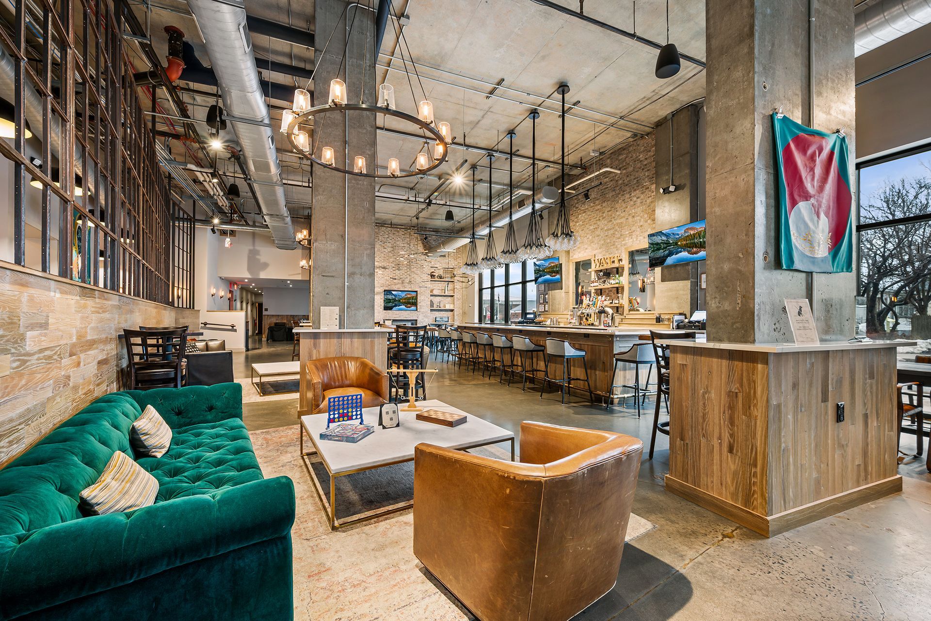 A lounge area with a green velvet sofa and leather armchairs, leading to a wooden bar counter in a modern, industrial room.