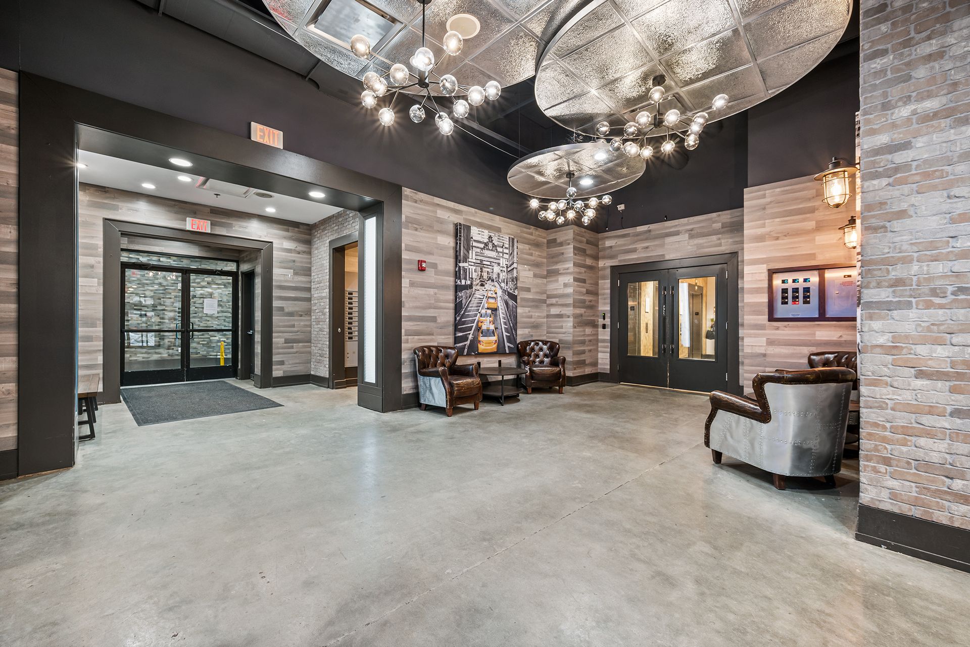 Modern industrial lobby with brick walls, concrete floors, metallic orb chandeliers, and brown leather armchairs.