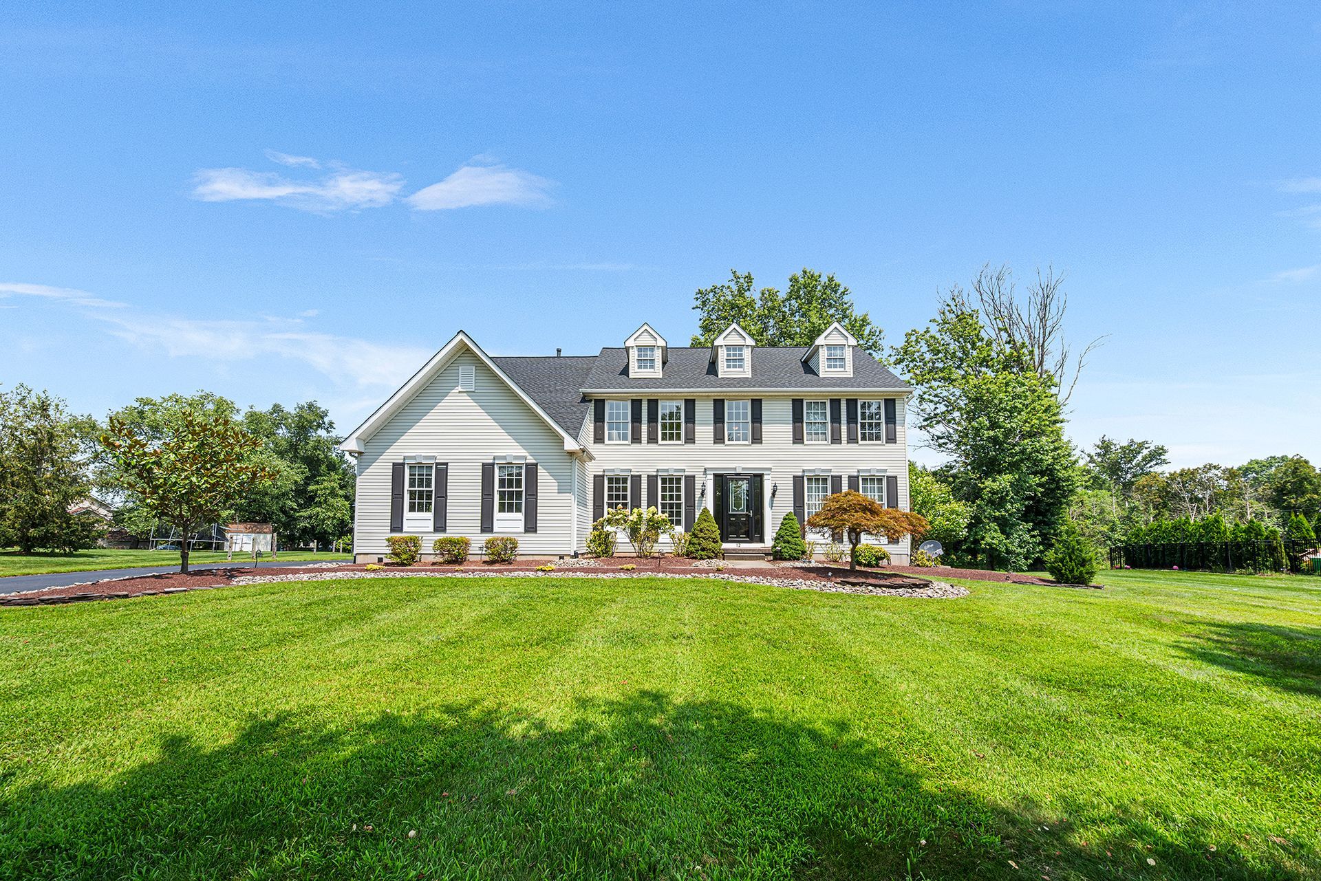 A light-colored two-story colonial style house with black shutters, set behind a large, sunlit grassy lawn under blue sky.