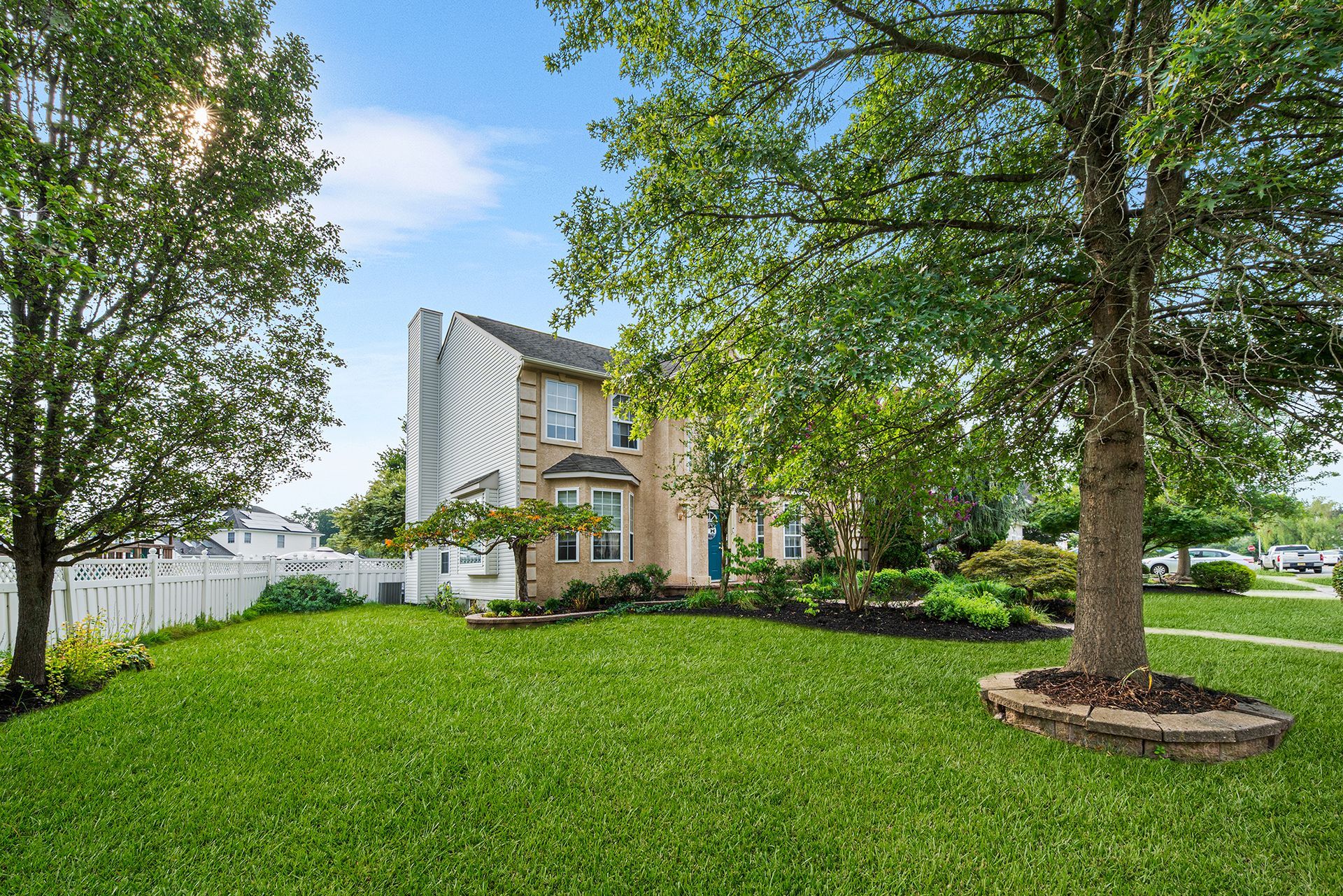 Two-story brick house with a blue front door, surrounded by green lawns and mature trees under a bright blue sky.