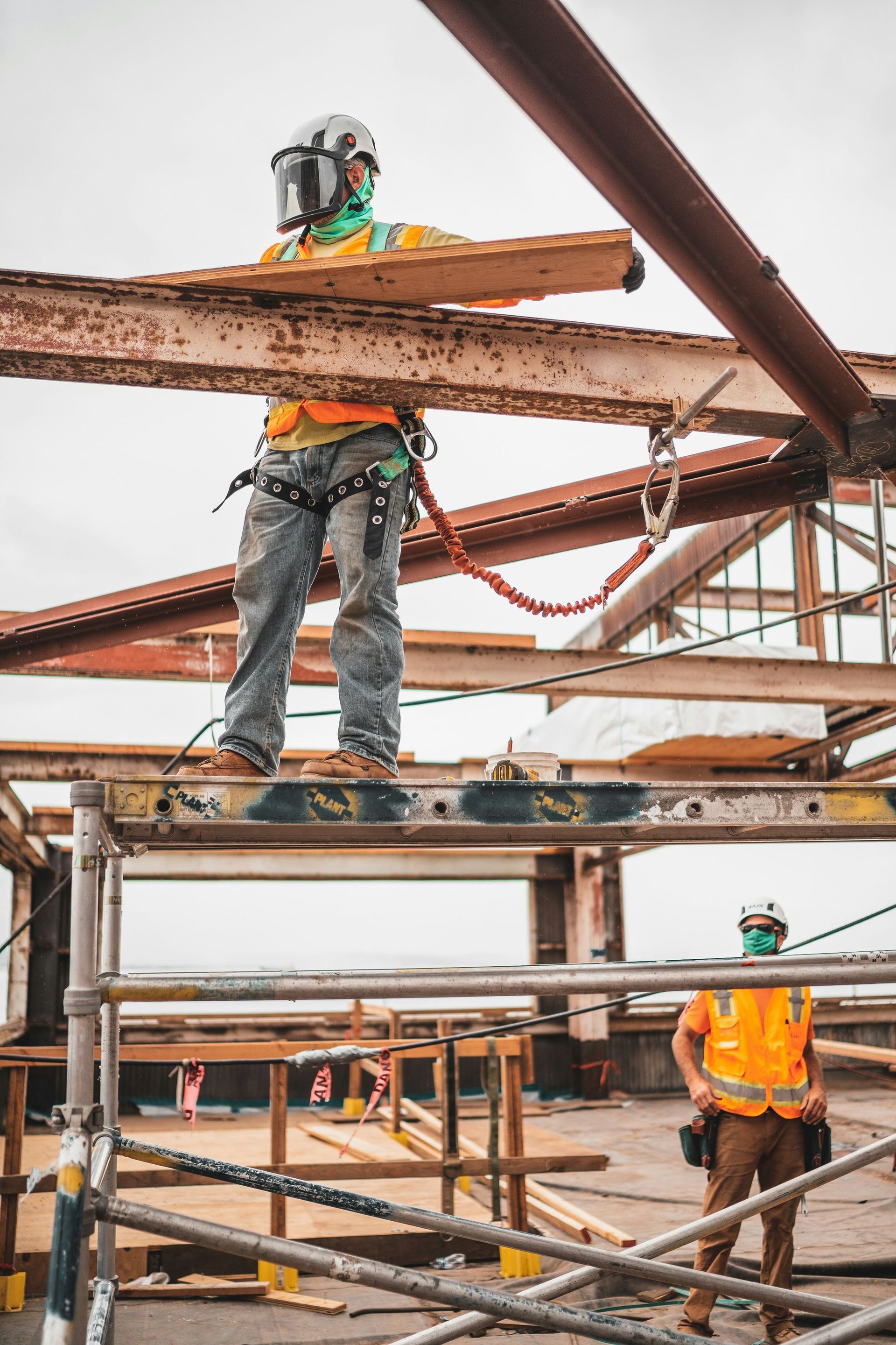 Two construction workers in safety gear and masks work on a steel frame structure.