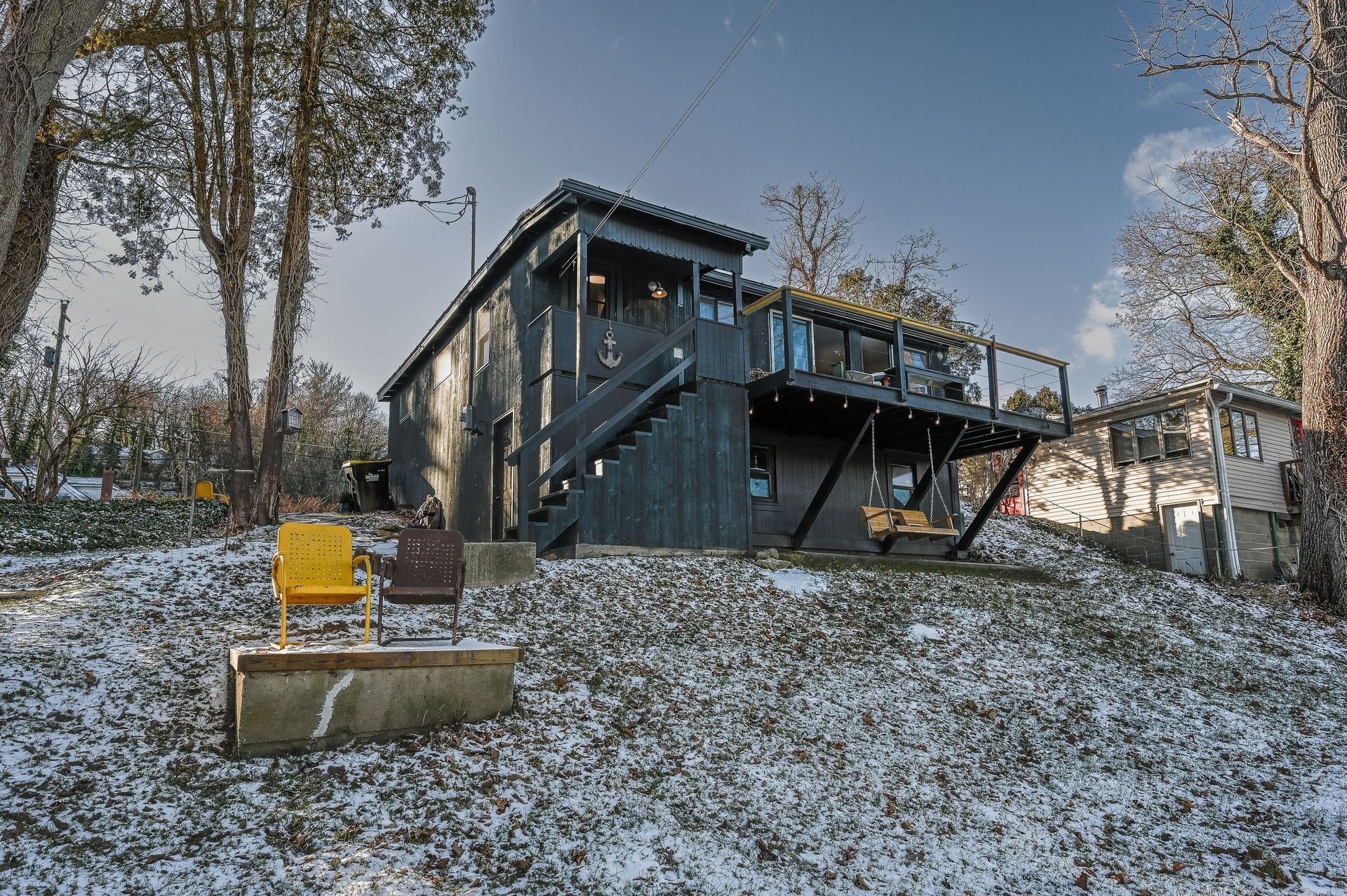 A dark-sided two-story house with a wooden deck, set on a snowy, tree-lined hill with two chairs on a concrete ledge.