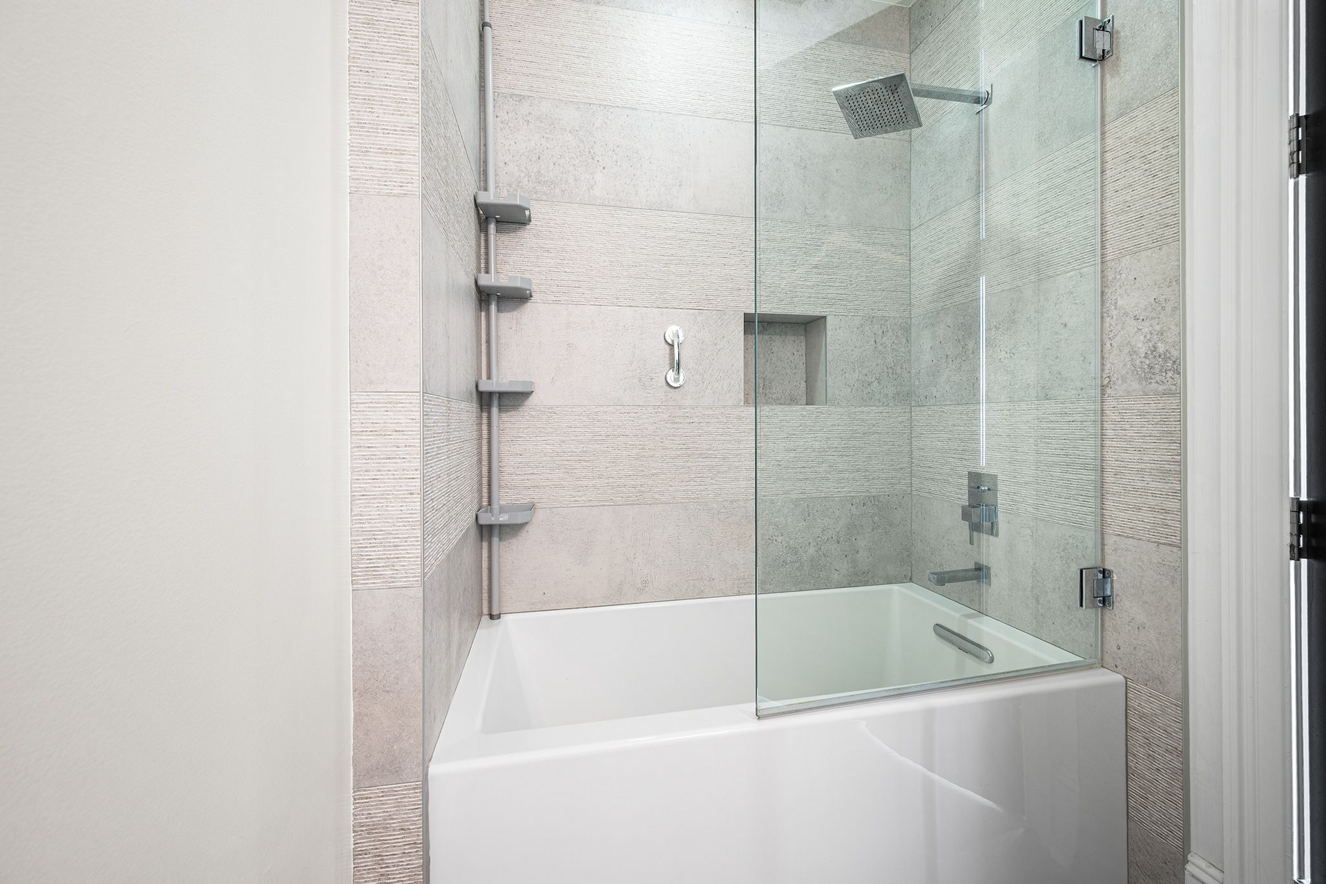 A white bathtub with a glass shower door and shelves on a wall with textured, light-colored stone tile.