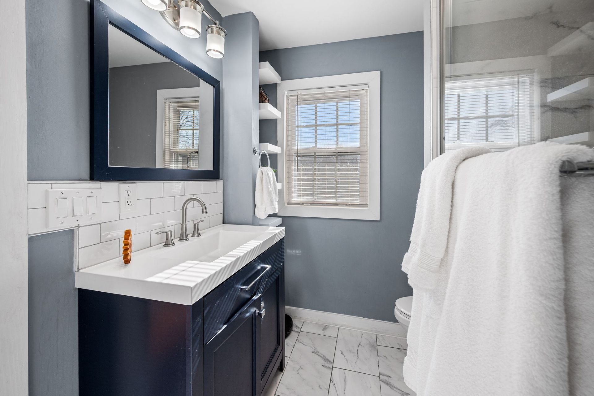 A blue vanity with a white countertop and rectangular mirror in a bathroom with grey walls and a white-tiled backsplash.