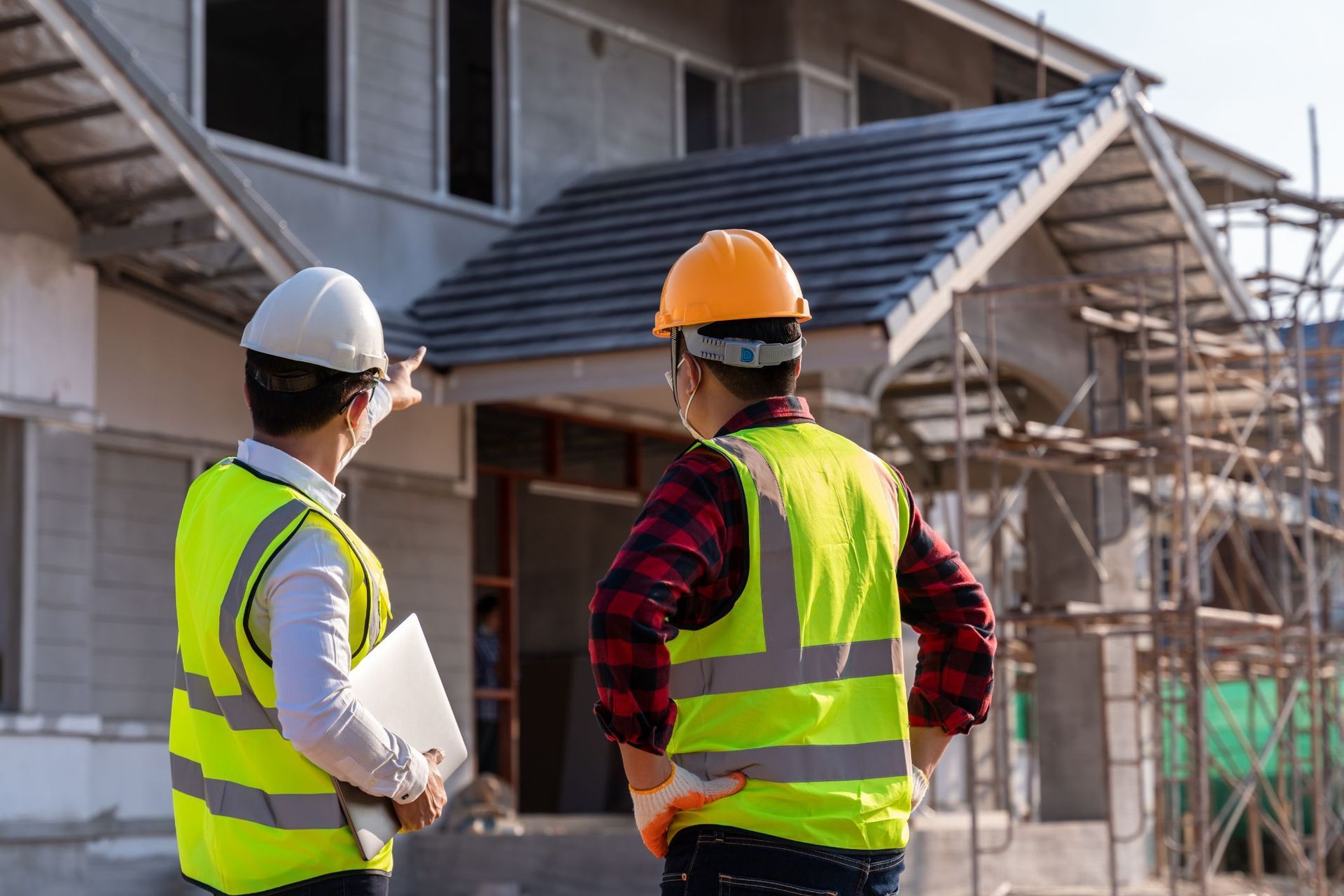 Two construction workers in safety vests and hard hats inspect a house under construction.