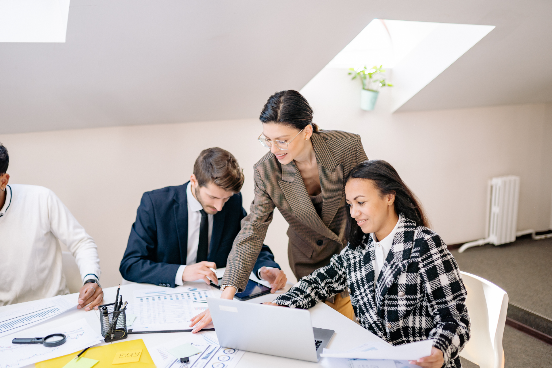 Coworkers collaborating around a laptop at a bright office table, reviewing documents and notes