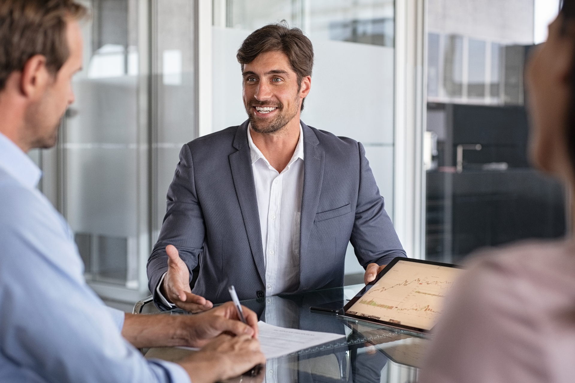 Business meeting in a glass-walled office, with a man in a blazer speaking across a conference table.