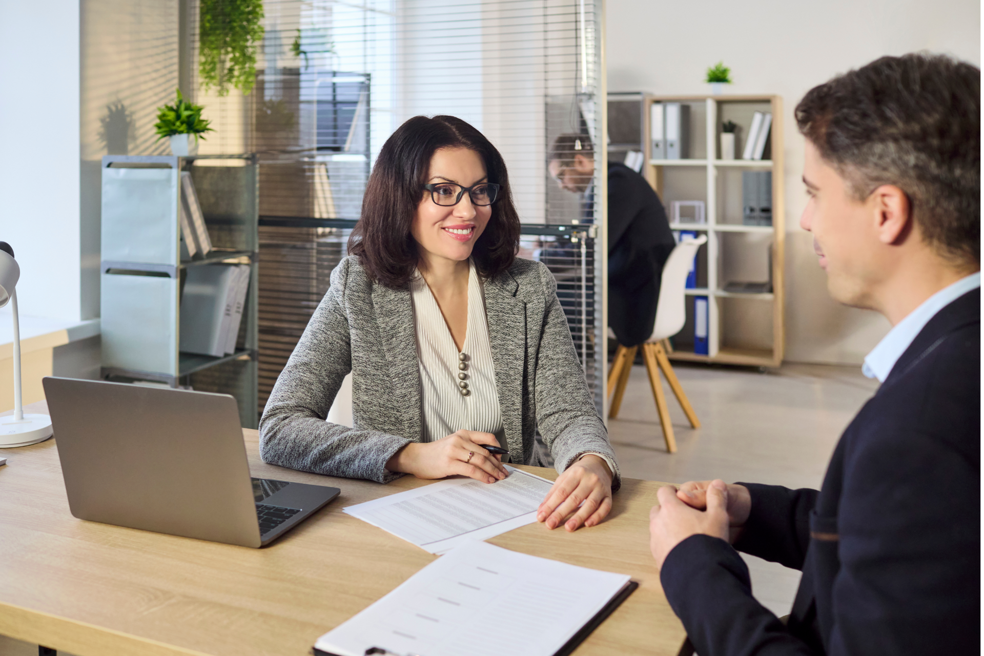 Two coworkers in a bright office discussing papers across a desk with a laptop open nearby