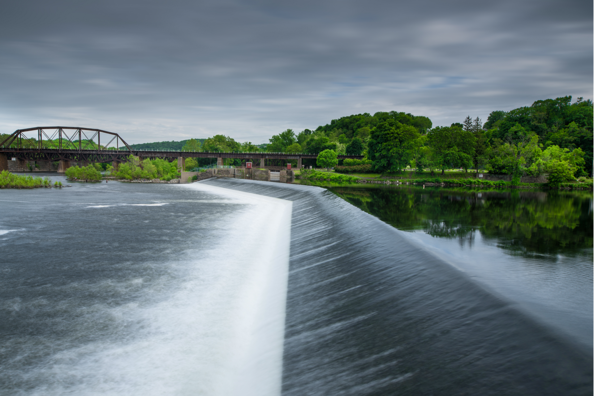 Wide river weir with white water flowing under an overcast sky, green trees and a bridge in the background
