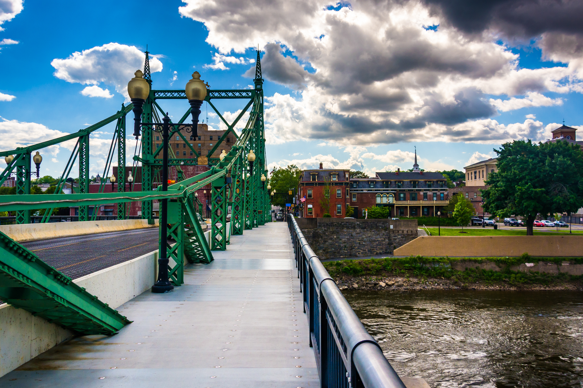 Green truss bridge over a river beside buildings under a cloudy blue sky
