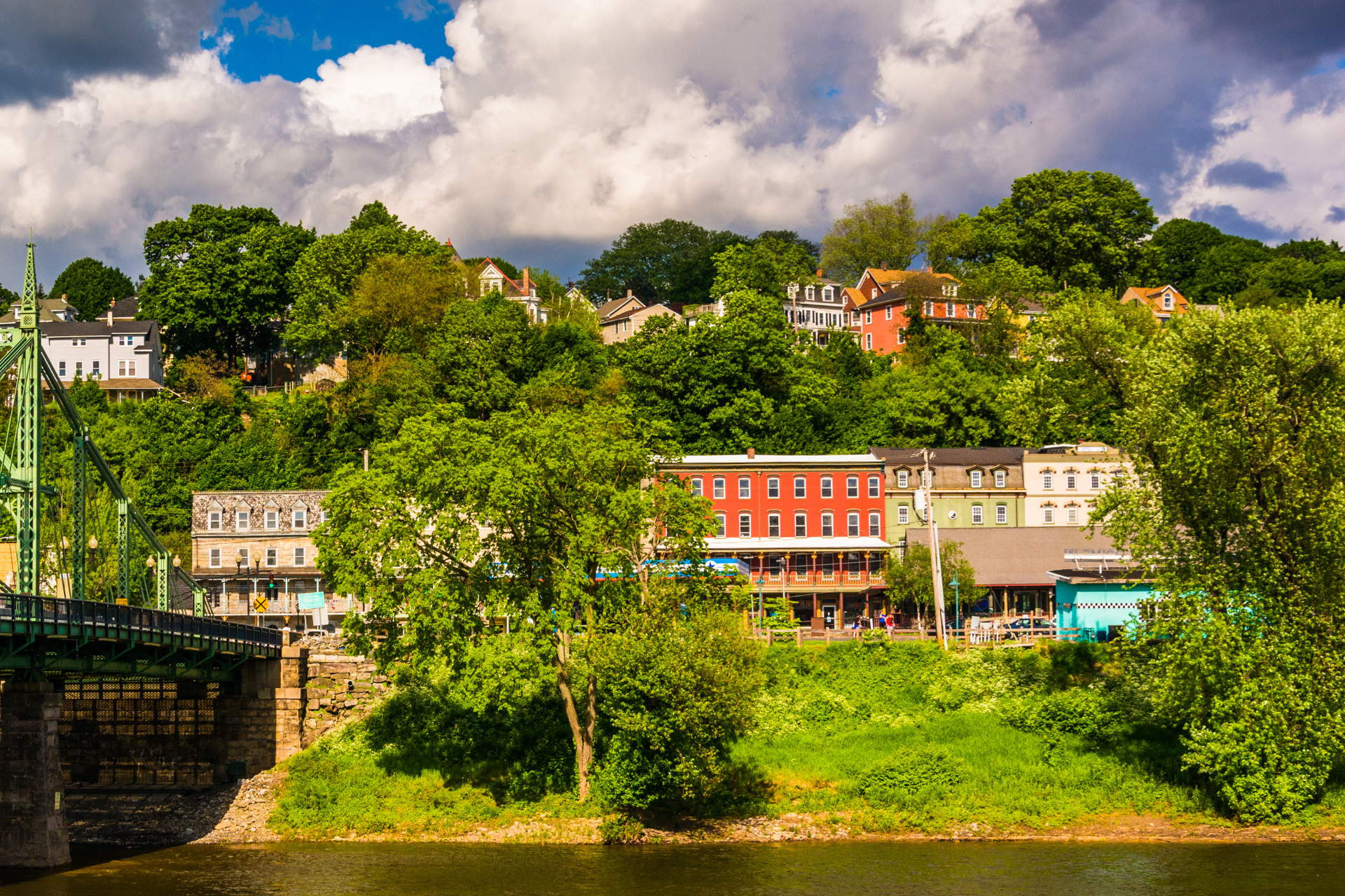 Colorful riverside village with houses, trees, and a bridge under a partly cloudy sky