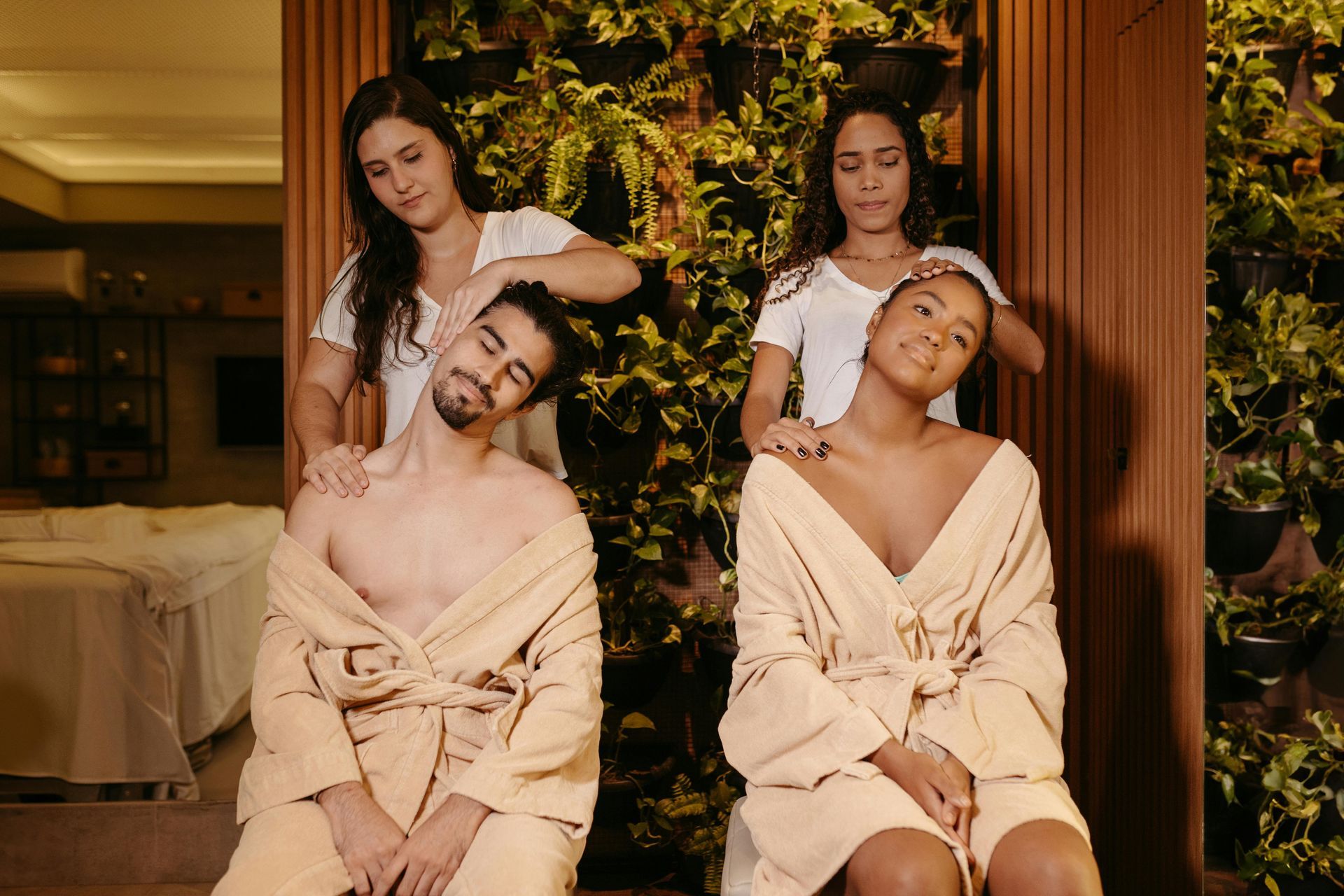 Four people in robes pose indoors, seated against a lush green wall in a warm-lit spa setting