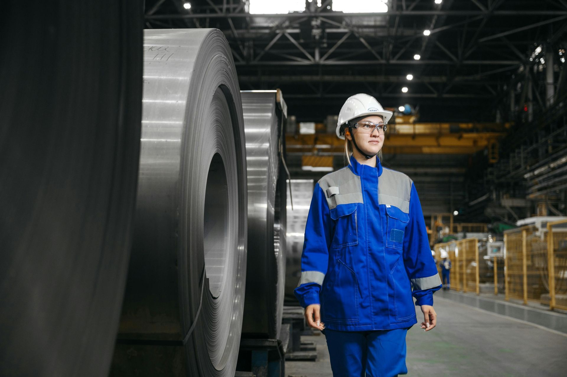 Worker in blue safety gear and hard hat walking beside large industrial equipment in a factory warehouse.