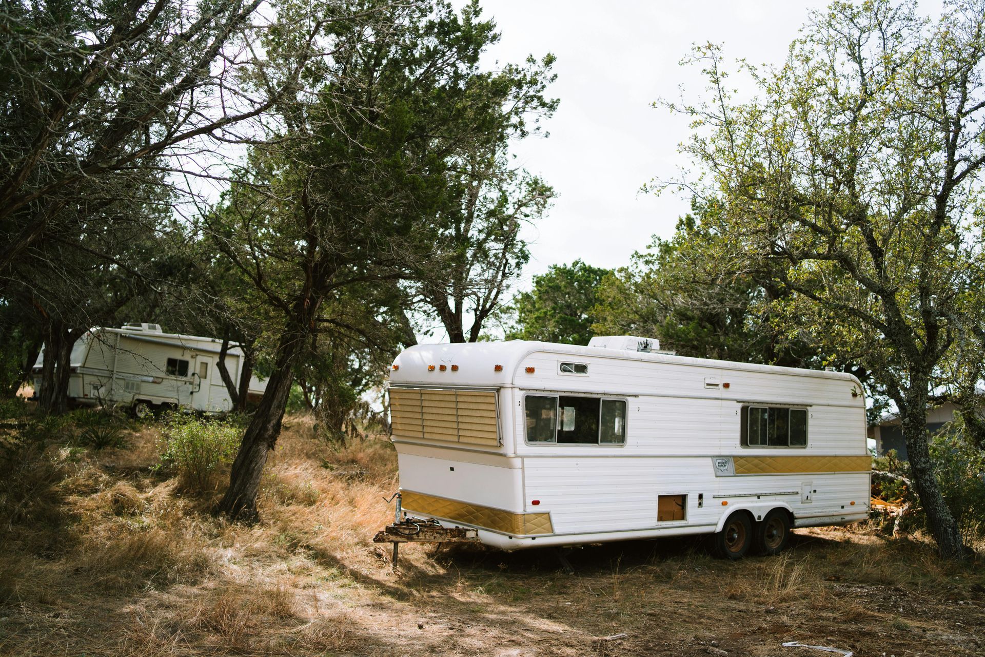 White and tan travel trailer parked in a wooded campsite with another camper in the background.