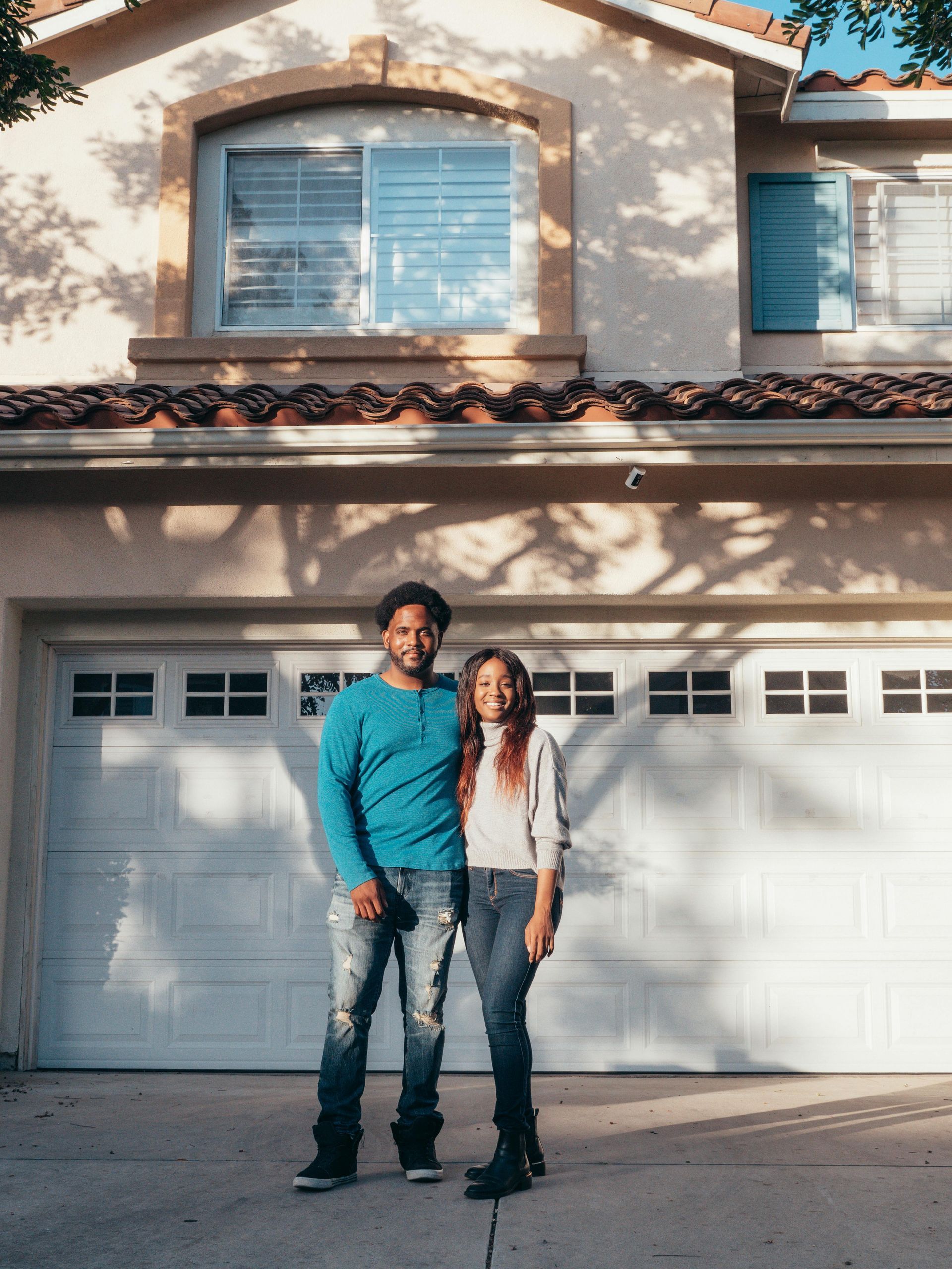 Two people standing in front of a house and garage, with beige siding and white doors.