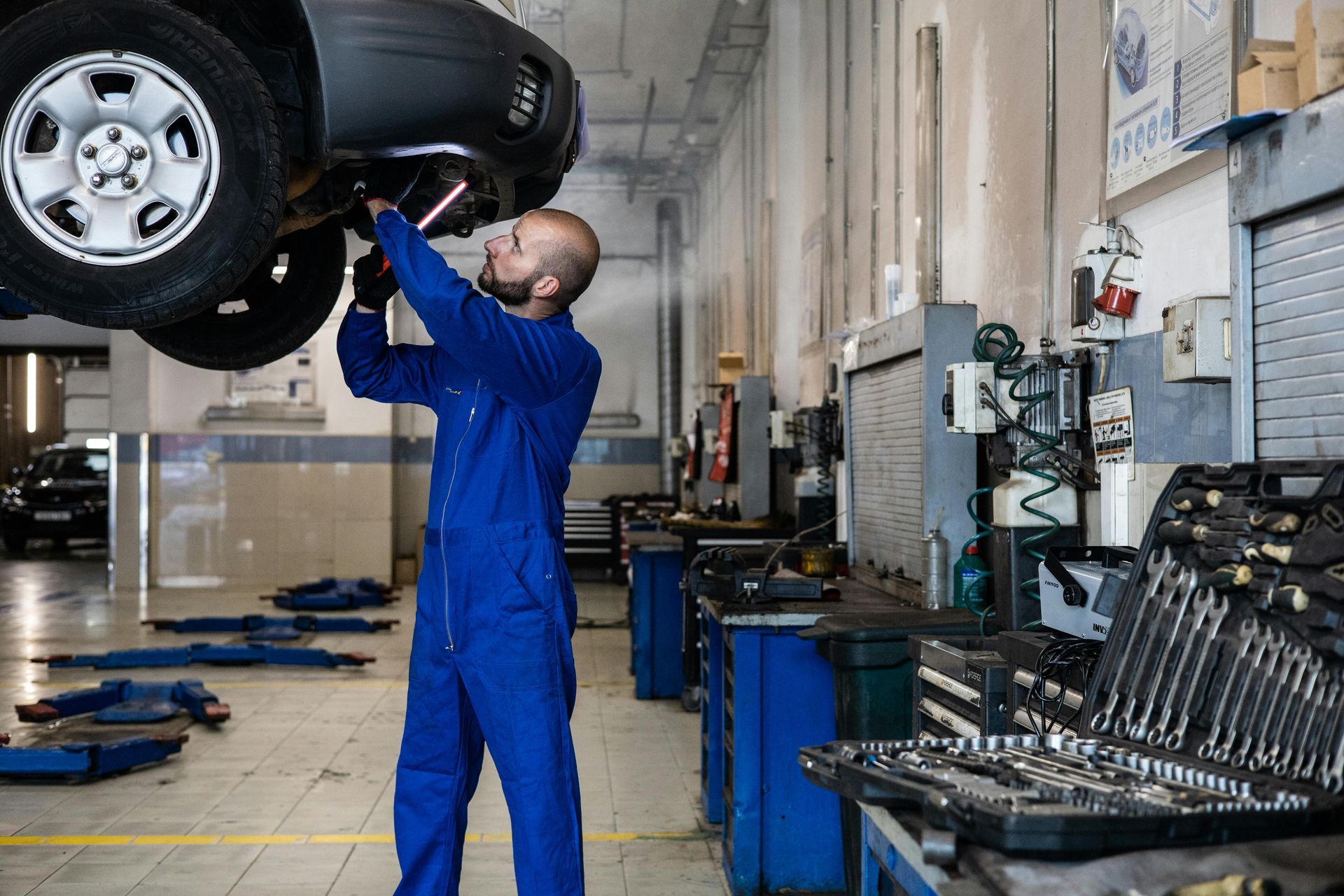 Mechanic in blue coveralls inspecting the underside of a lifted car in an auto repair shop.