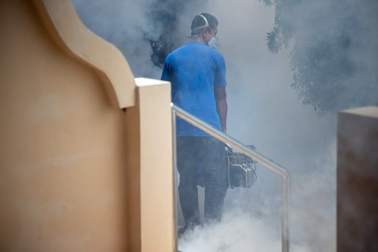 Person in blue walks through thick smoke carrying a tool near a building staircase