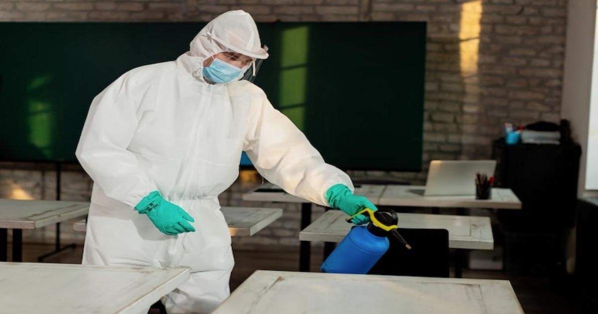 Worker in white hazmat suit spraying disinfectant onto tables in a room