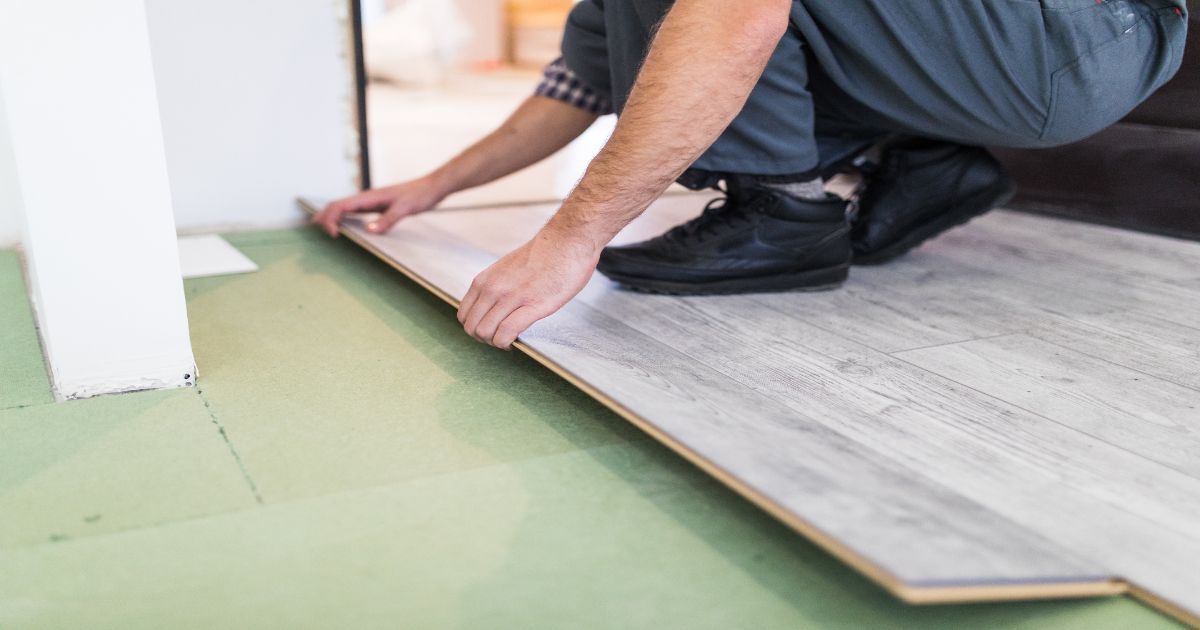 Person installing a light gray floorboard beside a white wall on a green underlayment