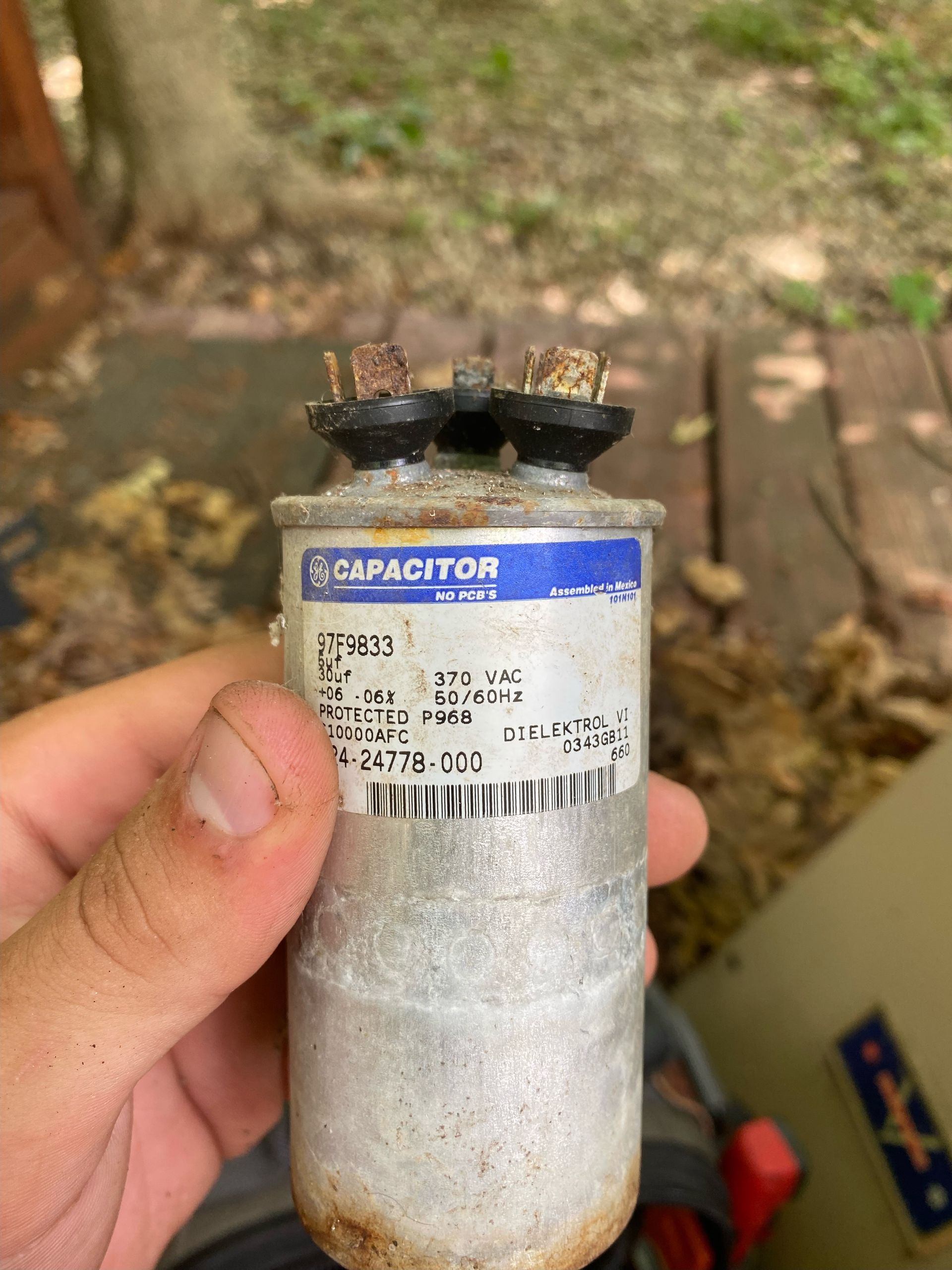 A person holds a rusted, cylindrical HVAC dual run capacitor with clearly visible electrical terminals and a label.