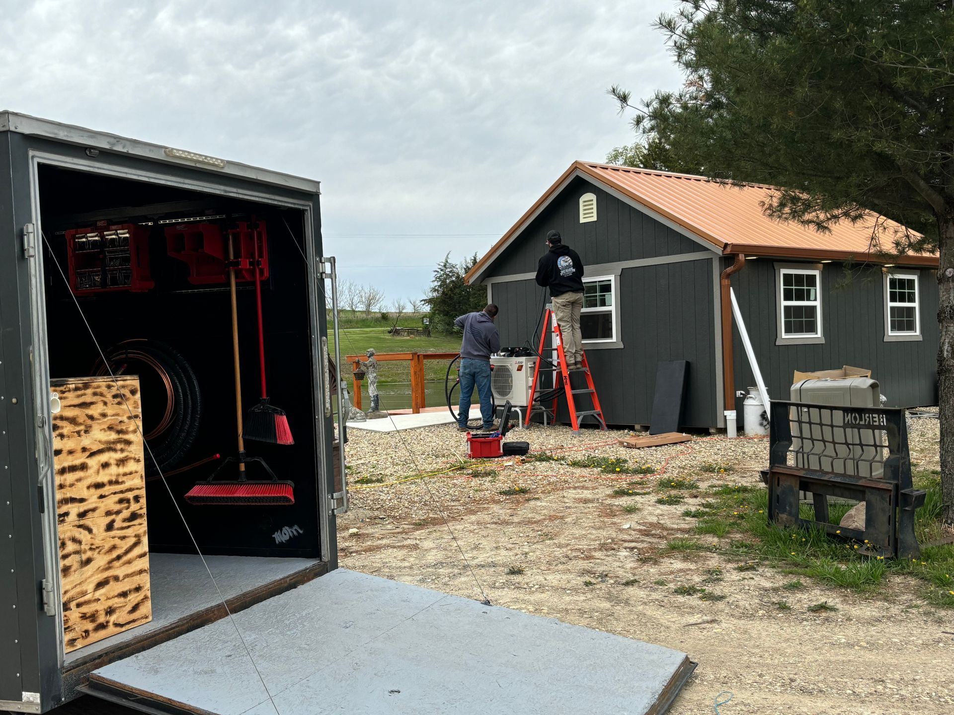 Two people perform maintenance on a small gray building, one on a ladder, with an open utility trailer in the foreground.