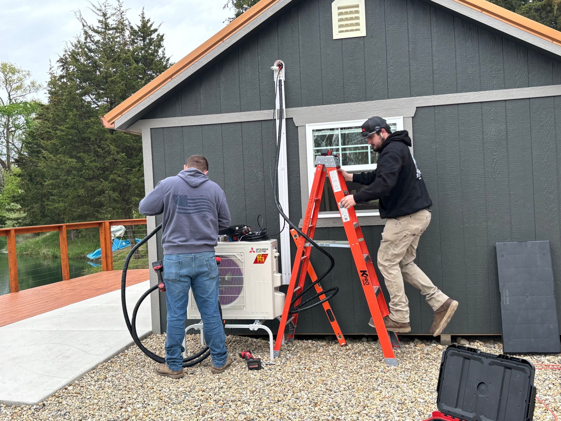 Two technicians install a mini-split HVAC unit on the exterior of a small grey shed.