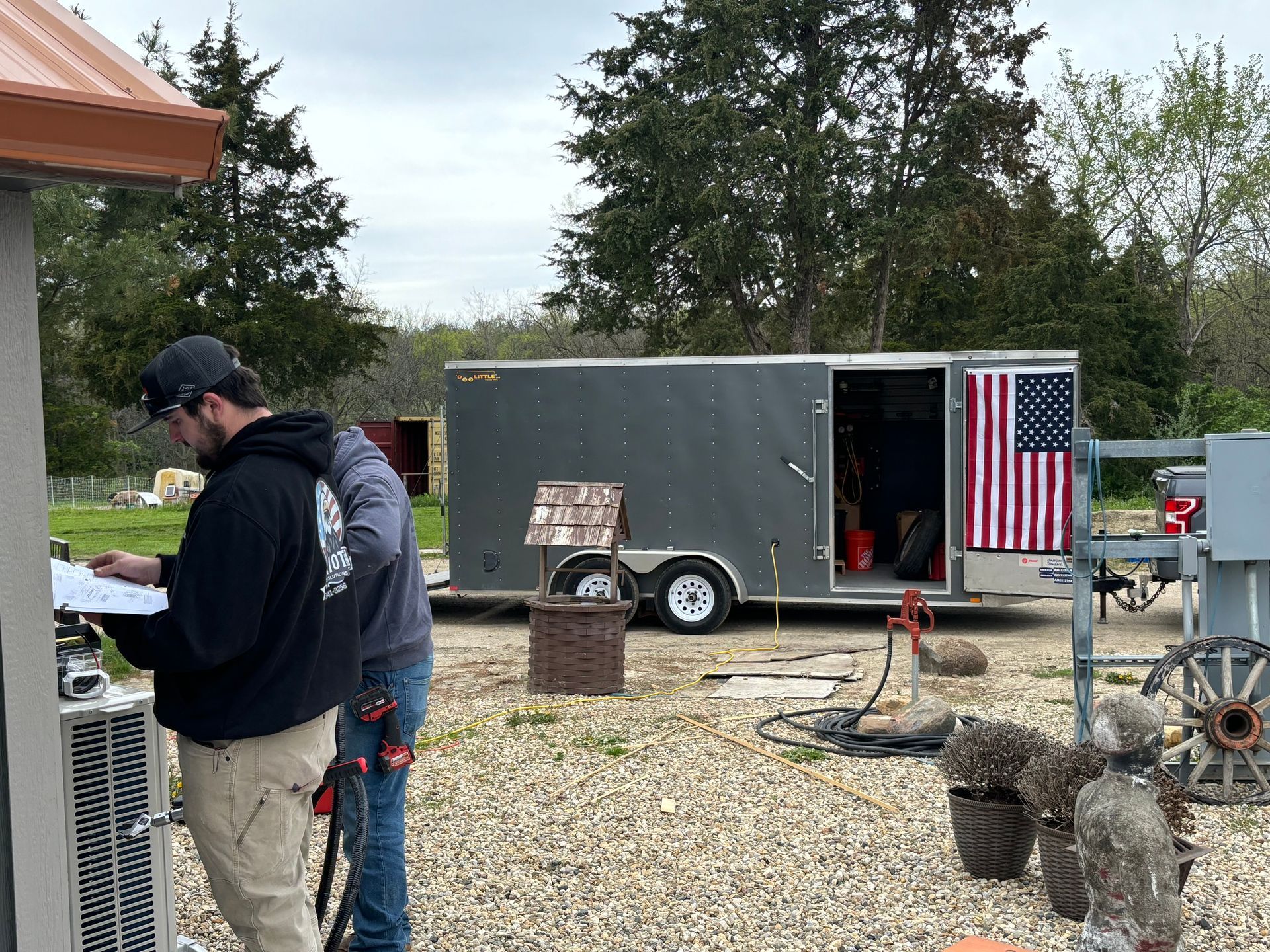 Two workers in hoodies stand outdoors, reviewing paperwork near a mechanical unit beside an open cargo trailer.