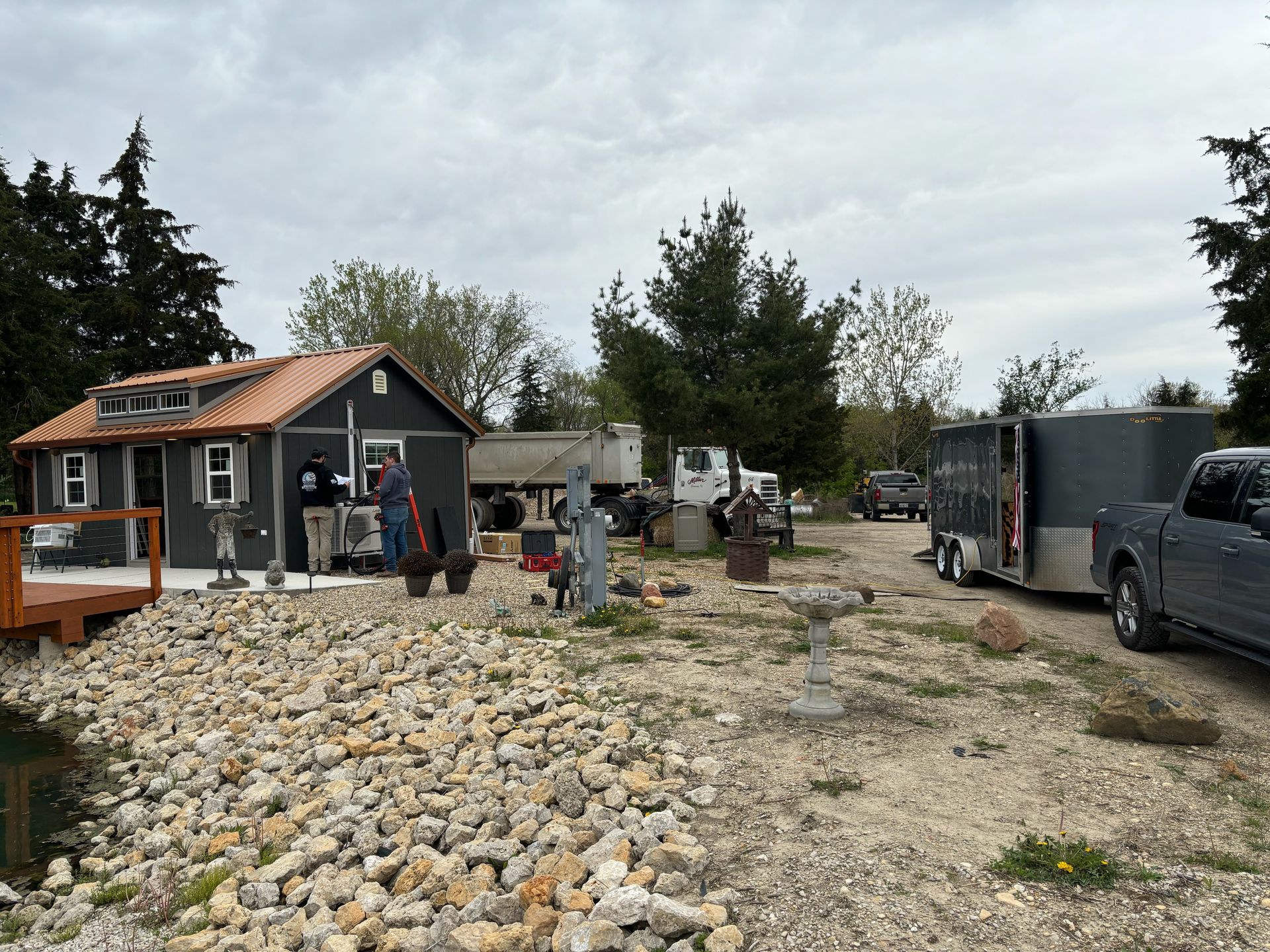 Two people work on a small dark-gray house near a rocky pond, with a dump truck and a truck-towed trailer parked nearby.